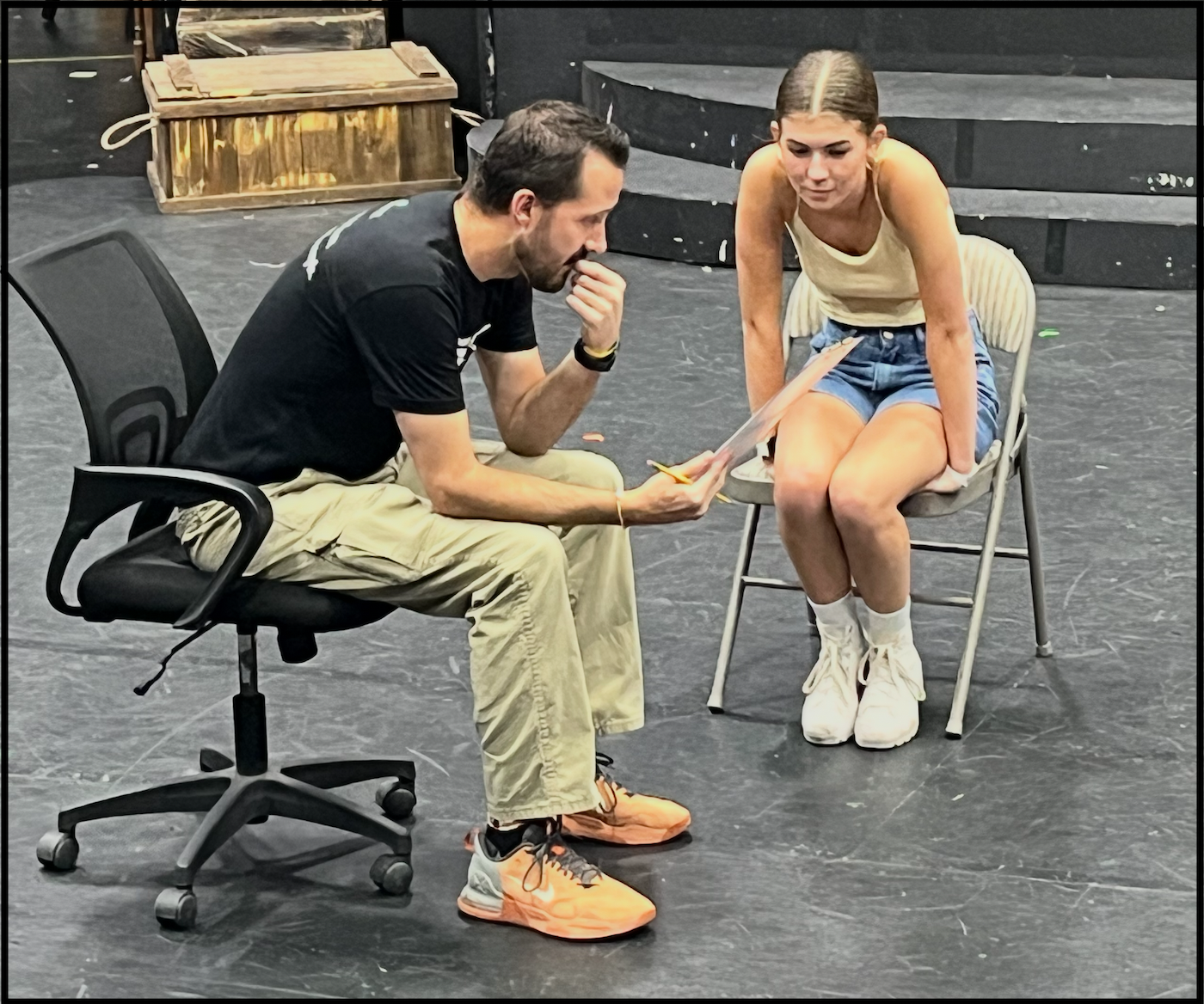 A man and a woman sitting on chairs in what appears to be a theater or rehearsal space, engaged in a discussion over a script or piece of paper.