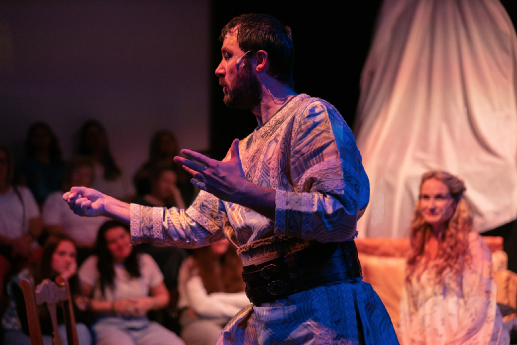 A man with dark hair and beard wearing a patterned shirt, standing on stage with his hands raised, performing in front of an audience. In the background, a woman with blonde hair, also in costume, is seated and smiling, with a theatrical set piece behind them.