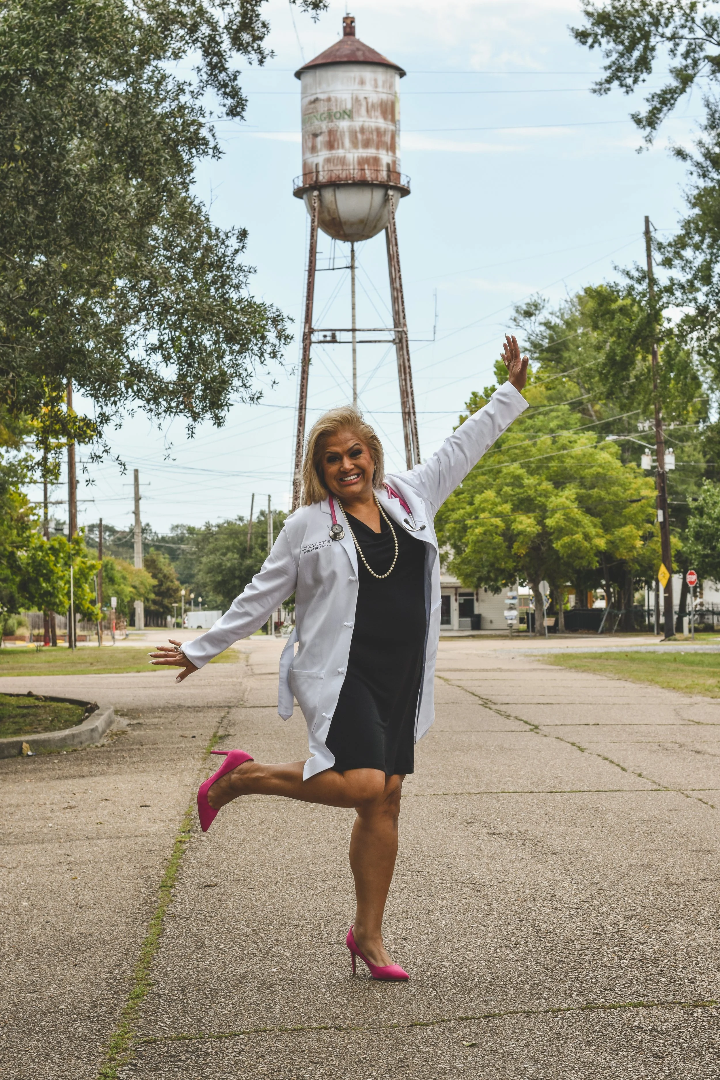A woman dressed as a doctor with a stethoscope smiling and jumping on a street with trees and a rusty water tower in the background.