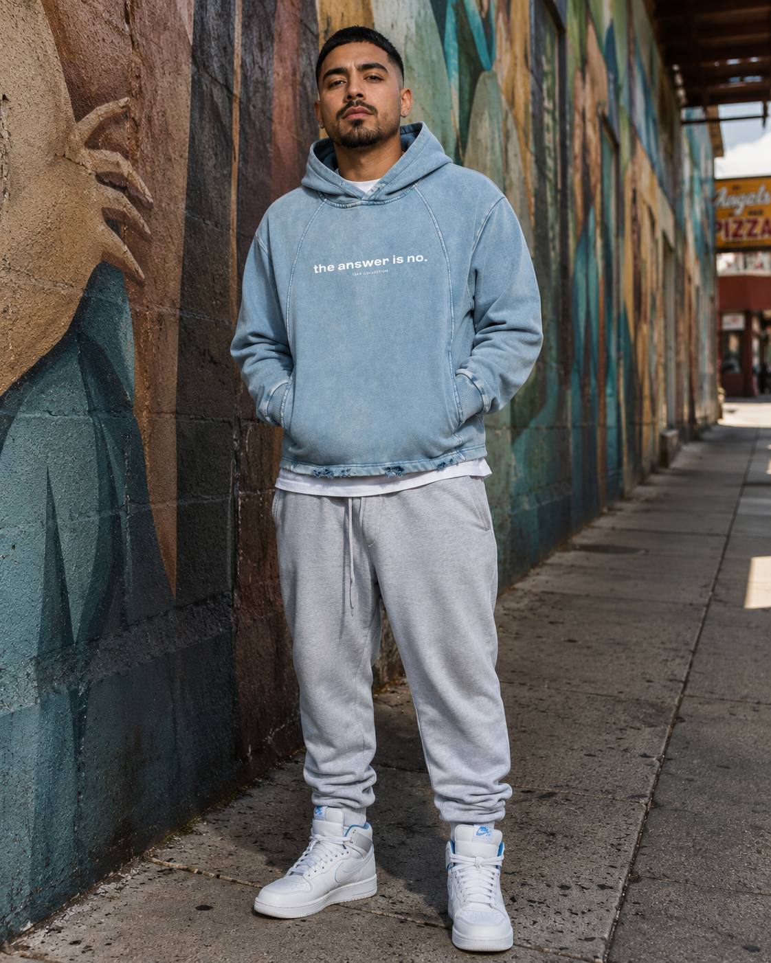 A man standing on a city sidewalk near a colorful mural wearing a light blue hoodie with the text "the answer is no." in white, gray sweatpants, and white Nike sneakers.