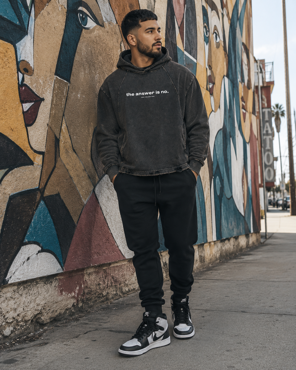A young man with dark hair and a beard stands with hands in pockets, wearing a black hoodie with white text that reads 'the answer is no.' over a colorful mural of various faces painted on a wall, with a city street and palm trees in the background.