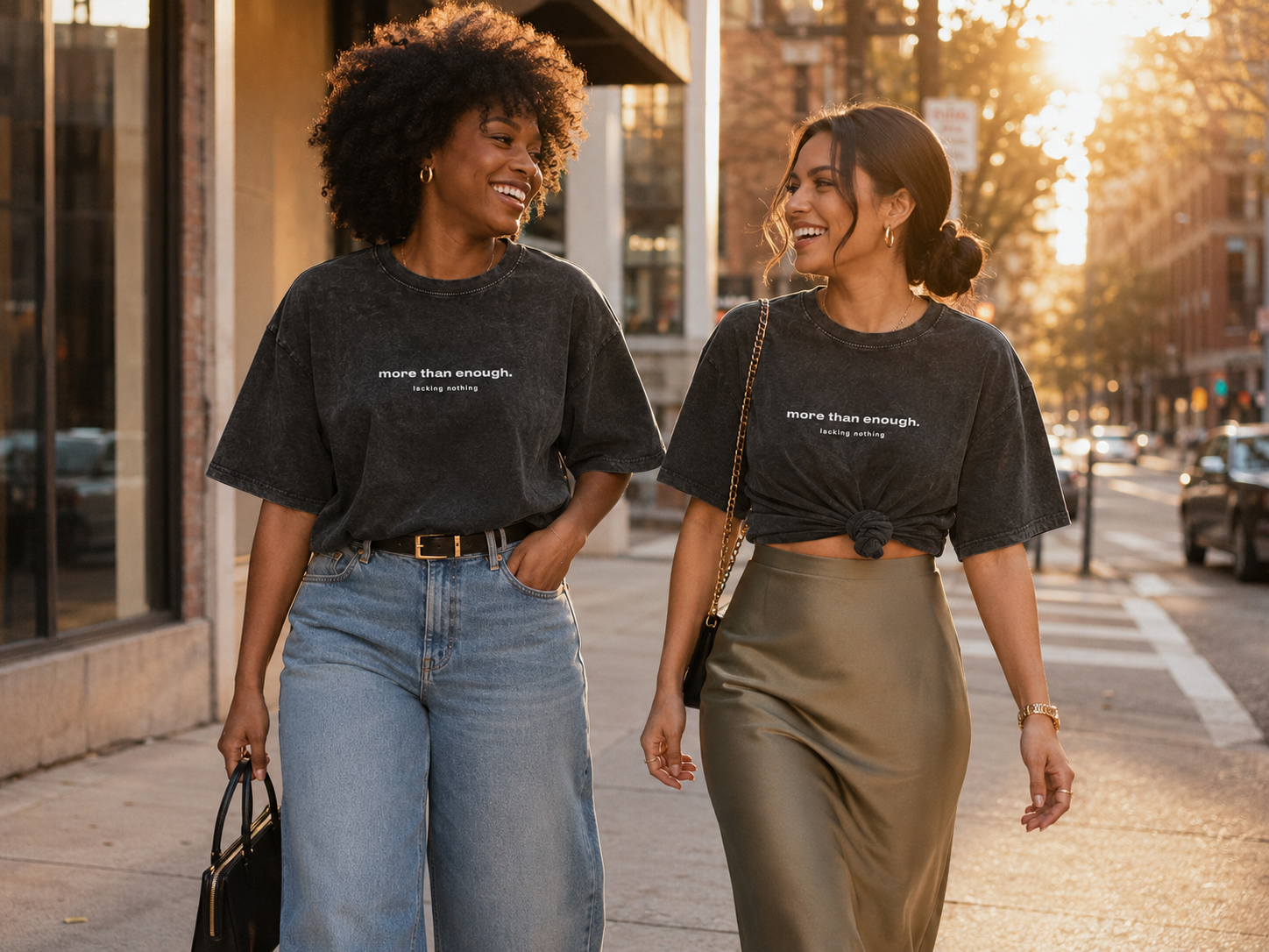 Two women walking on a city sidewalk at sunset, smiling and looking at each other, wearing matching black t-shirts with the phrase 'more than enough. lacking nothing.'