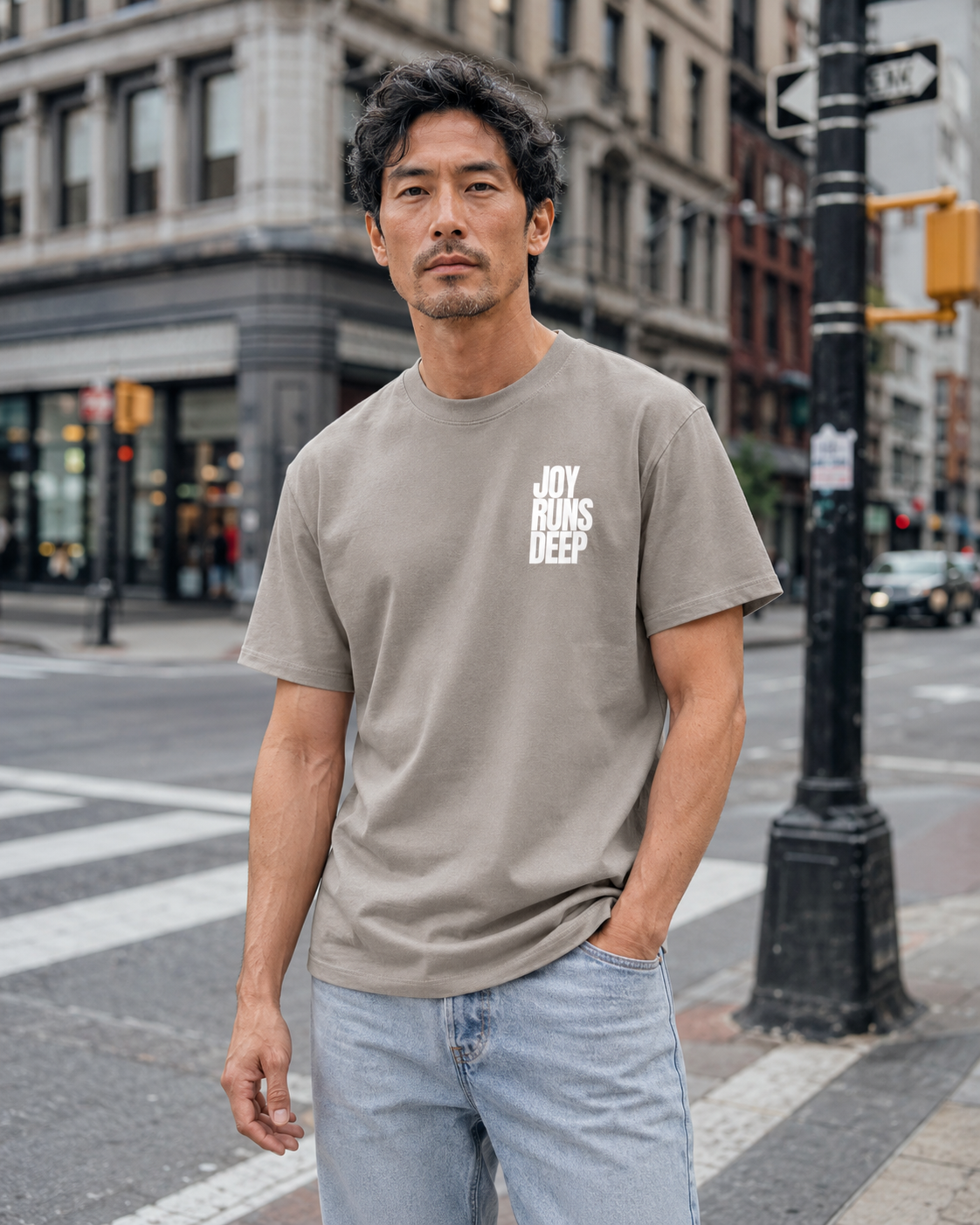 A man standing on a city street corner, wearing a gray T-shirt with the words "Joy Runs Deep" printed on it, and light blue jeans, with buildings and traffic signals in the background.