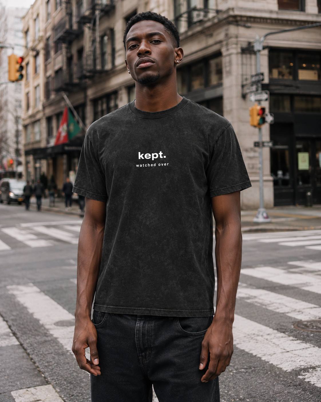 A young man standing at a crosswalk on a city street, wearing a black T-shirt with the words 'kept. watched over' printed on it, and black jeans, with buildings and traffic lights in the background.