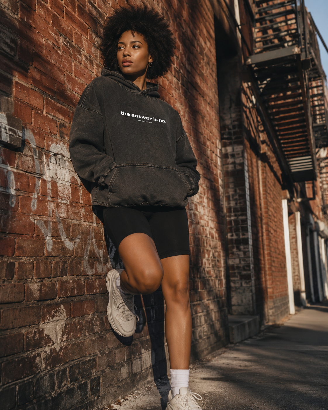 A young woman with curly hair leaning against a brick wall outdoors during daytime. She is wearing a black hoodie with the words 'the answer is no.' and black shorts, white sneakers, and white socks. She appears to be relaxed and looking off into the