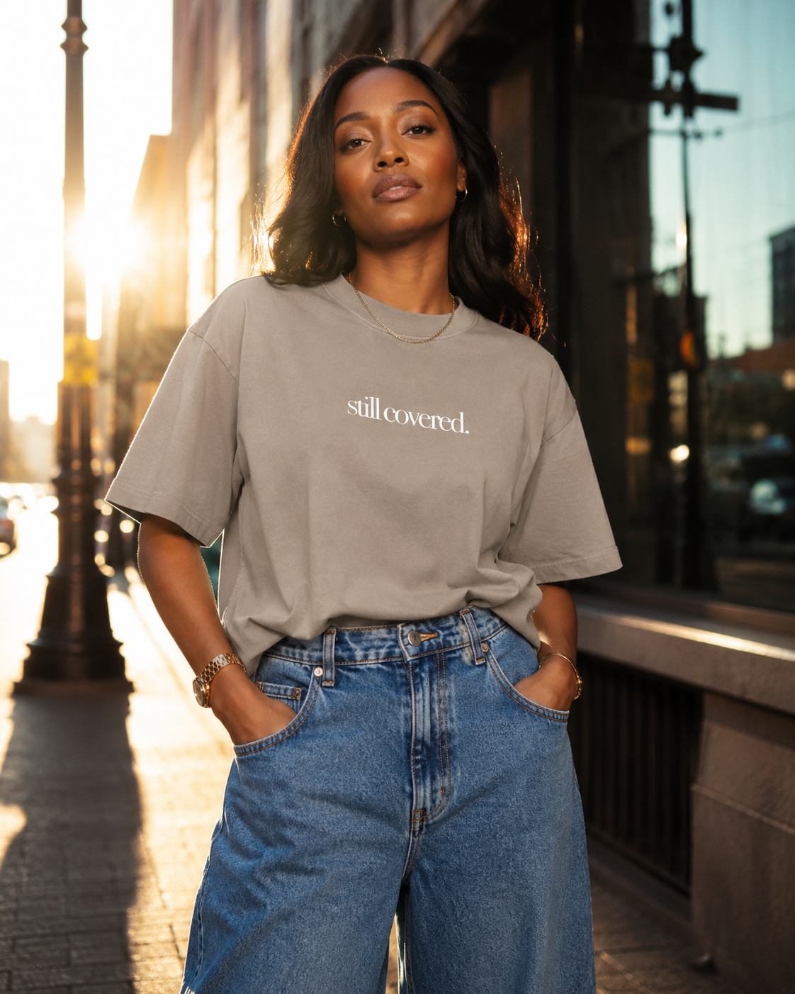 A woman in sunglasses and casual clothing stands on a city sidewalk during sunset. She wears a gray t-shirt with the phrase 'still covered.' and blue jeans, with her hands in her pockets.