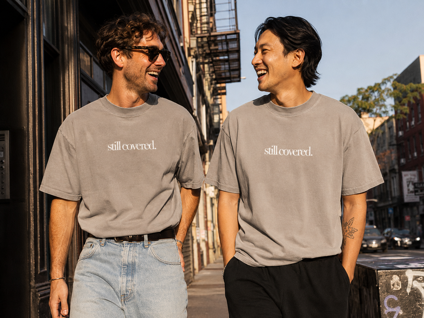 Two men walking and talking on a city sidewalk, wearing matching gray T-shirts that say 'still covered.' and smiling at each other.