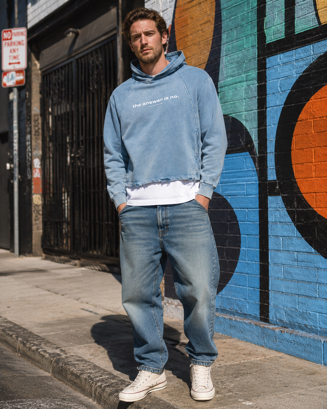 A young man with curly brown hair and a beard standing on a sidewalk in front of a colorful mural wall. He is wearing a light blue cropped hoodie with ripped hem and the text 'the answer is no.' on it, a white t-shirt underneath, baggy jeans, and whi