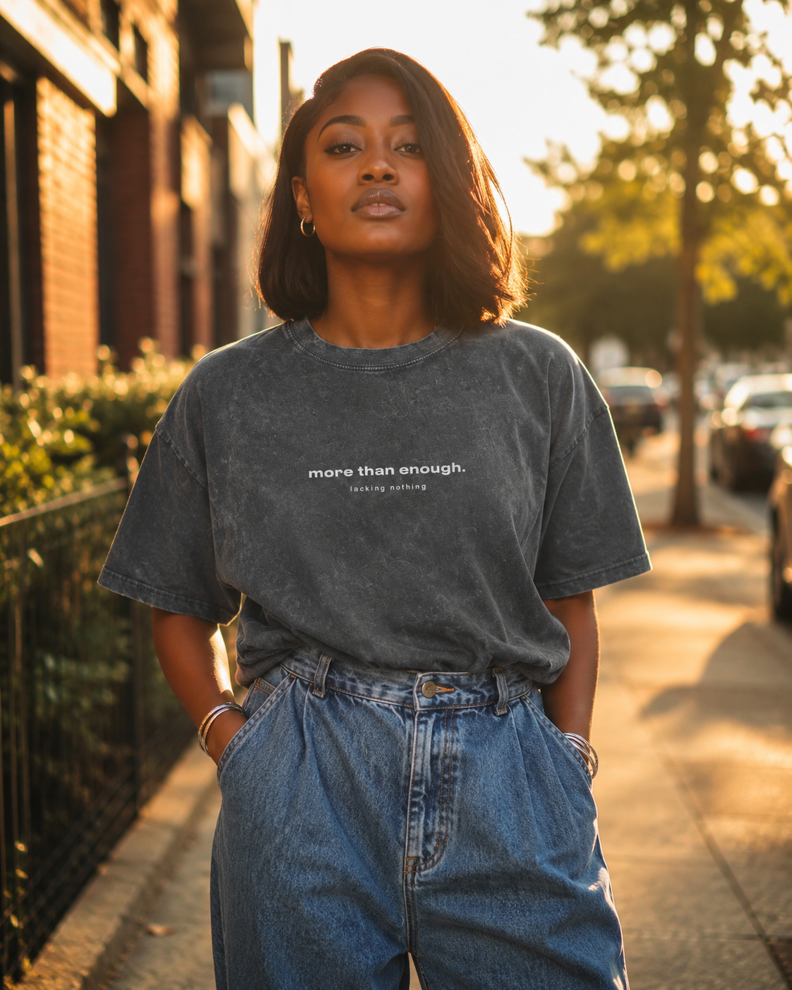 A young woman standing on a sidewalk during golden hour, wearing a black t-shirt with the text "more than enough. lacking nothing" and high-waisted blue jeans. She has shoulder-length dark hair, hoop earrings, and a confident expression.