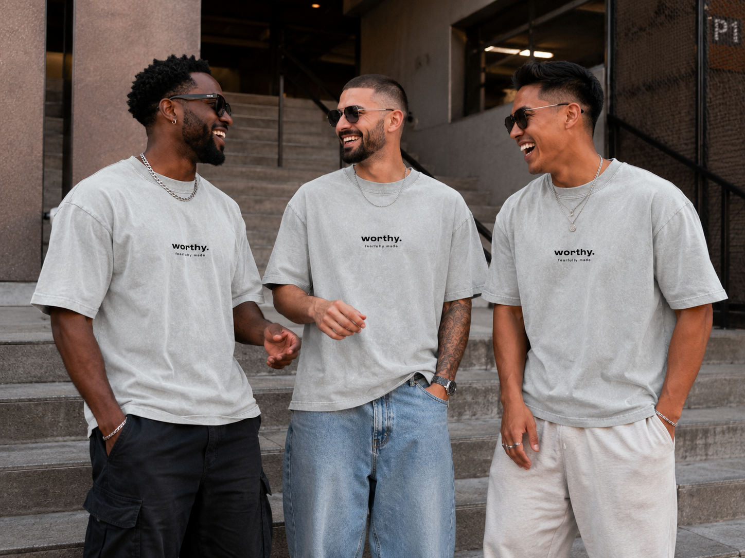 Three men wearing matching gray T-shirts and sunglasses are standing on steps outside, laughing and talking.