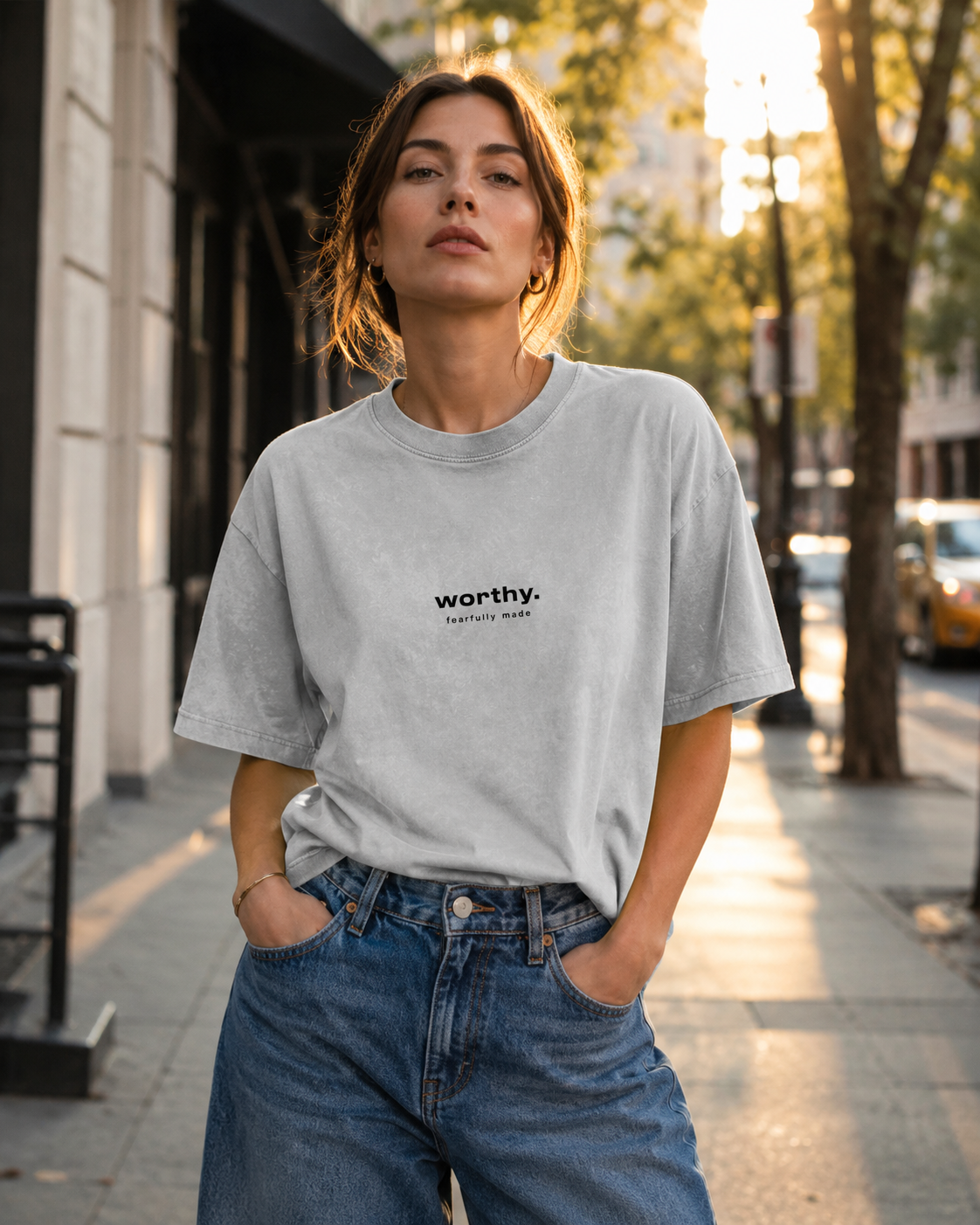 A woman standing on a city street in the late afternoon, wearing a light gray t-shirt with the words 'worthy. fearfully made' printed on it, and blue jeans, with trees and buildings in the background.