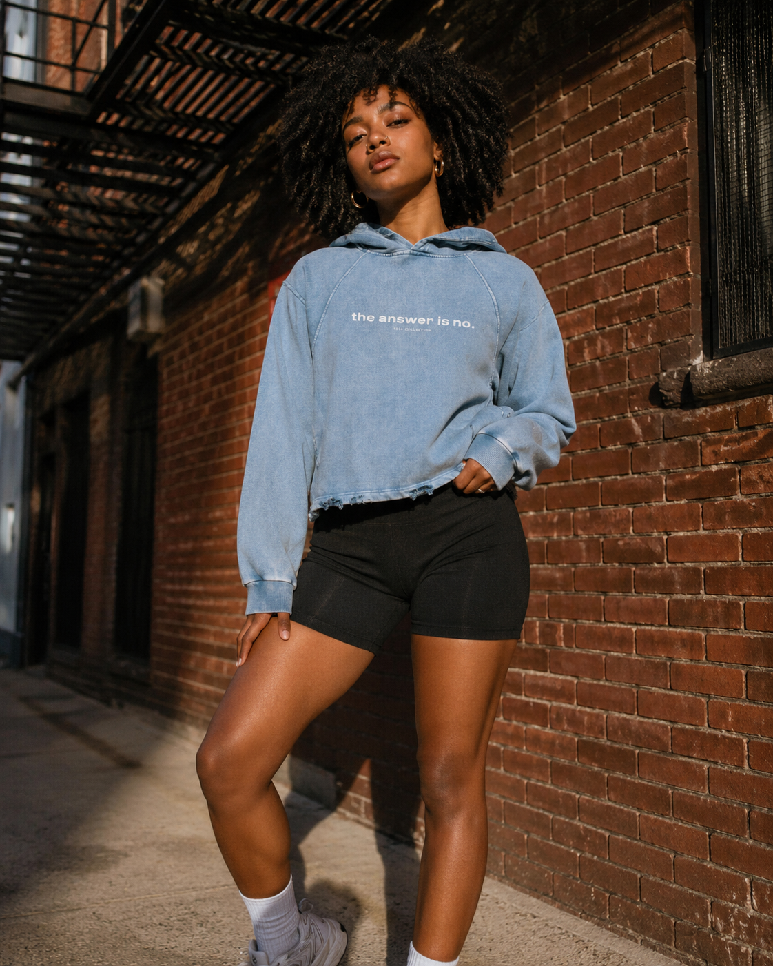 A young woman with curly hair standing in front of a brick wall, wearing a light blue cropped hoodie with the text "the answer is no." and black shorts, with sunlight casting shadows on the ground.