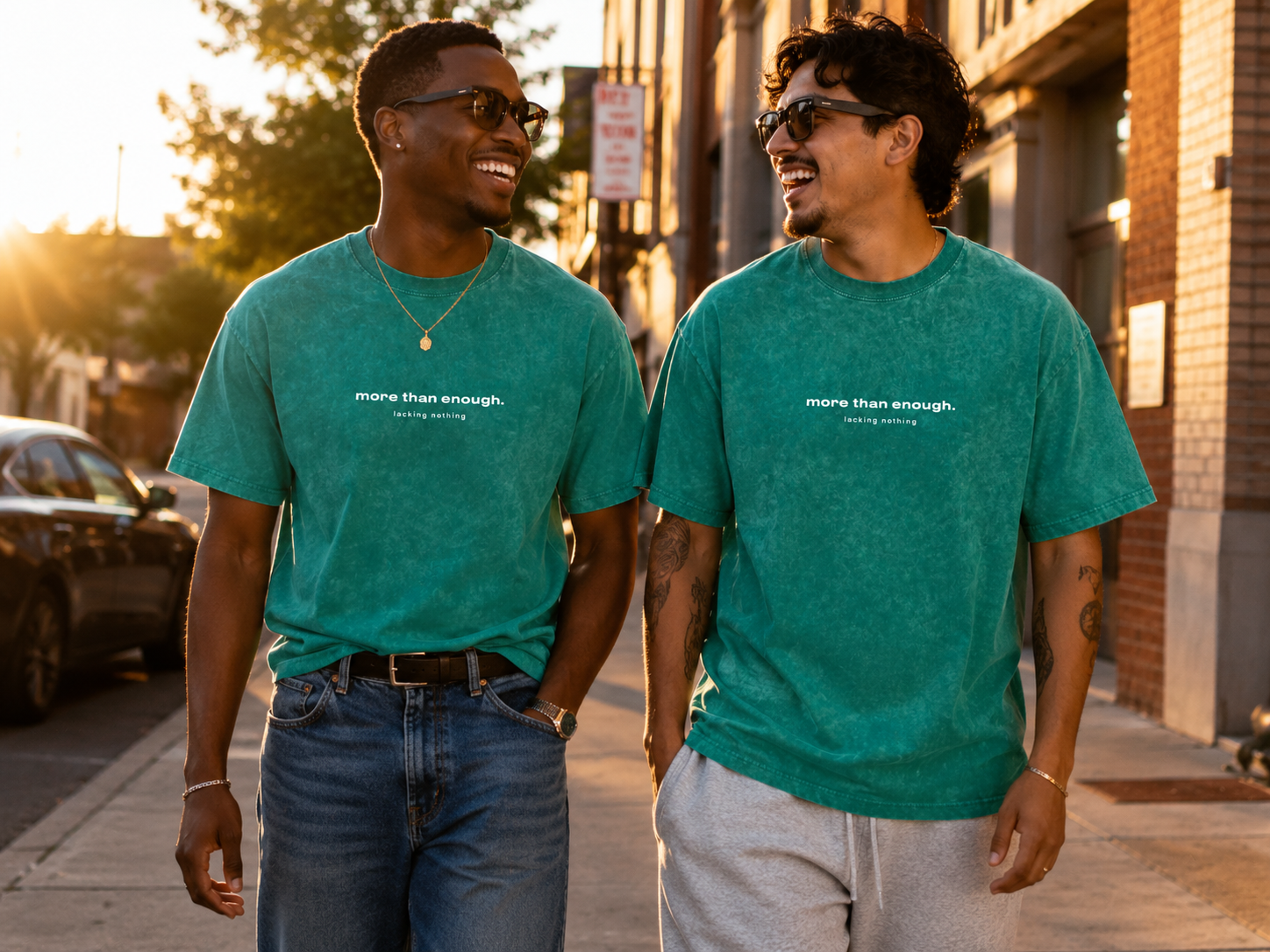 Two young men walking and talking on the sidewalk during sunset, both wearing teal t-shirts with the words "more than enough. lacking nothing," sunglasses, and casual jeans and shorts.