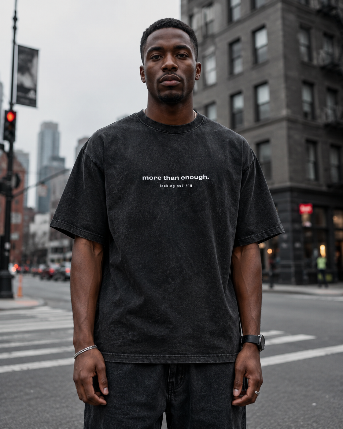 A young man standing on a city street wearing a black t-shirt that says 'more than enough. lacking nothing' with urban buildings and traffic lights in the background.