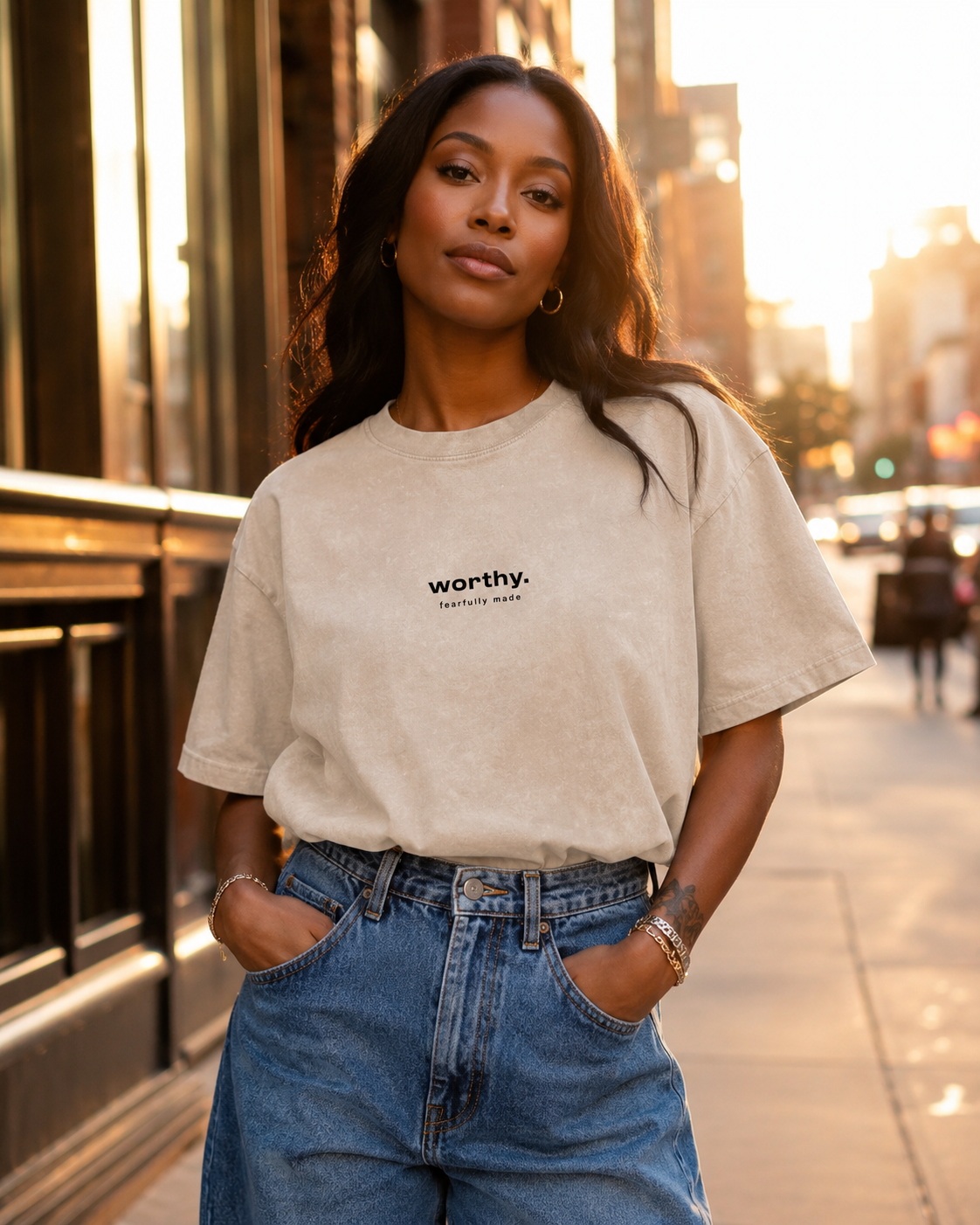 A woman standing outdoors on a city street during sunset, wearing a beige T-shirt with the word "worthy" printed on it and high-waisted blue jeans.