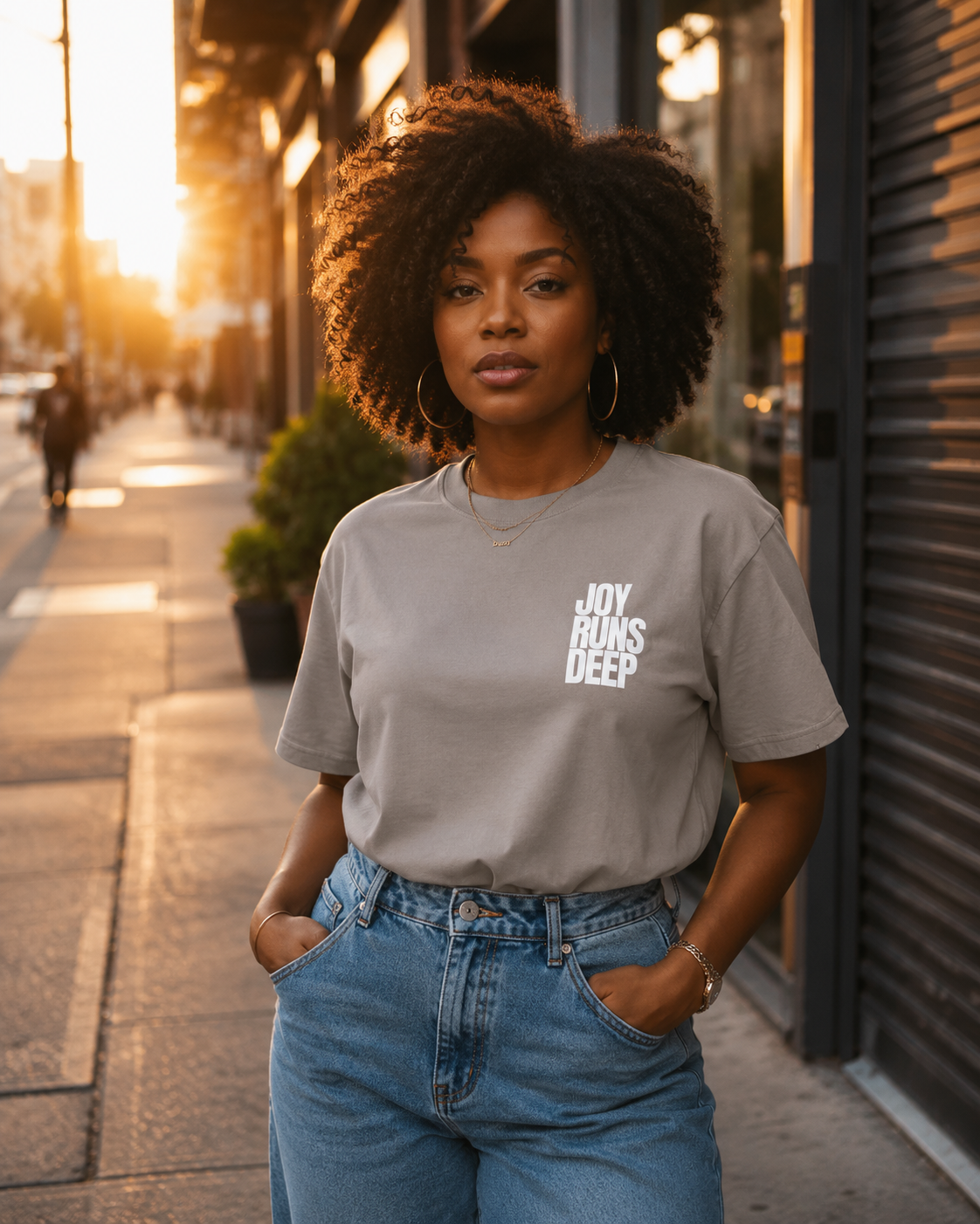 A young woman with curly hair standing on a city sidewalk during sunset, wearing a gray t-shirt with the text 'Joy Runs Deep' and blue jeans, with her hands in her pockets.
