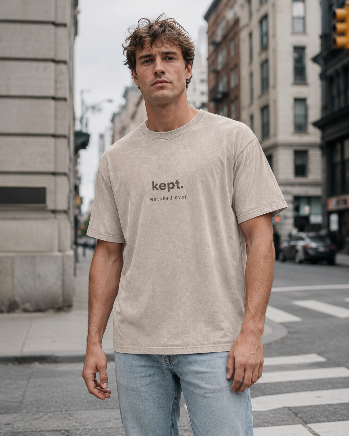 A young man standing on a city street corner wearing a beige T-shirt with the word 'kept.' and the phrase 'watched over' printed on it, with buildings and cars in the background.
