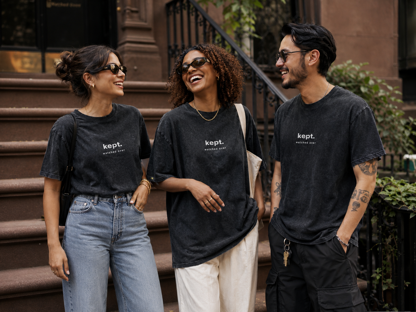 Three young adults standing outdoors on brownstone steps, engaged in conversation and smiling. All are wearing black t-shirts with the word 'kept.' and the phrase 'watched over' printed on them.