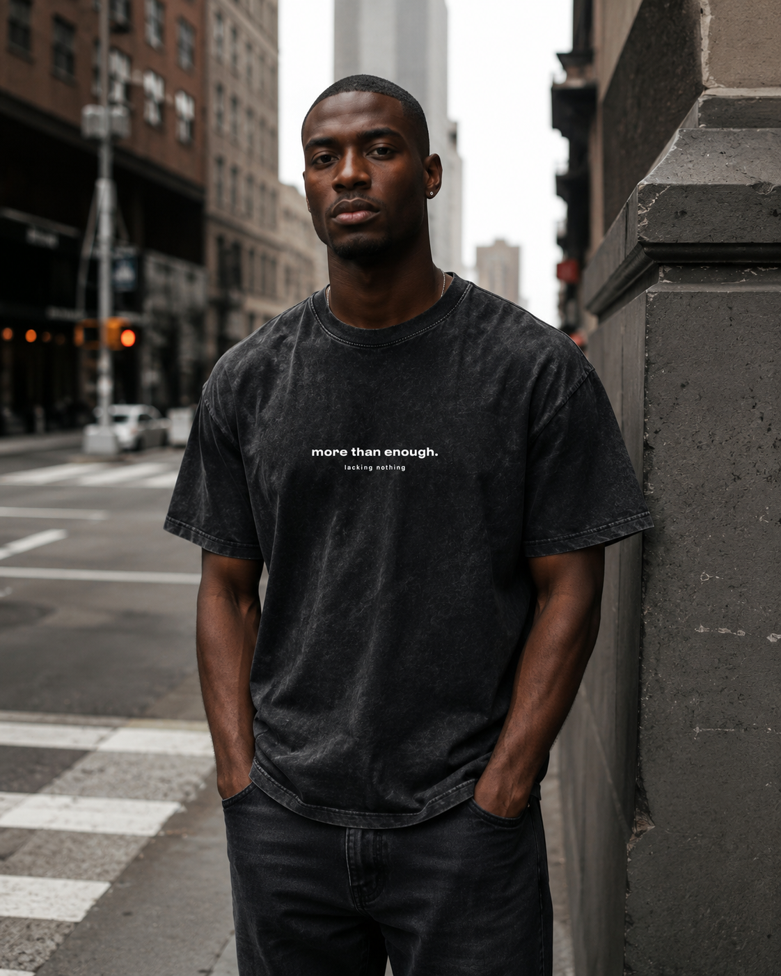 A young man with dark skin and short black hair stands on a city street corner, leaning against a gray building. He wears a black, slightly faded T-shirt with the words 'more than enough. lacking nothing' printed on it. The background features city b