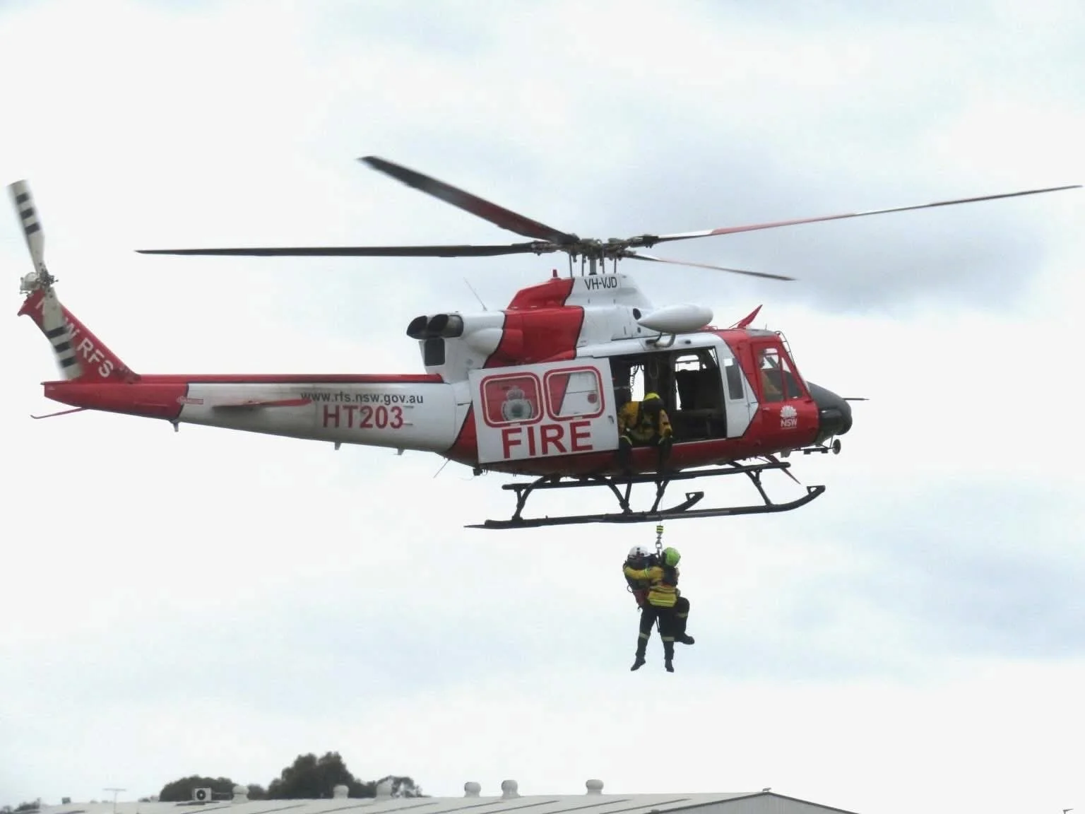A search and rescue helicopter in the sky, with a rescuer hanging from a harness below, assisting someone in a yellow vest. The helicopter is red and white, marked with fire department insignia and website, with open side doors and two personnel inside.