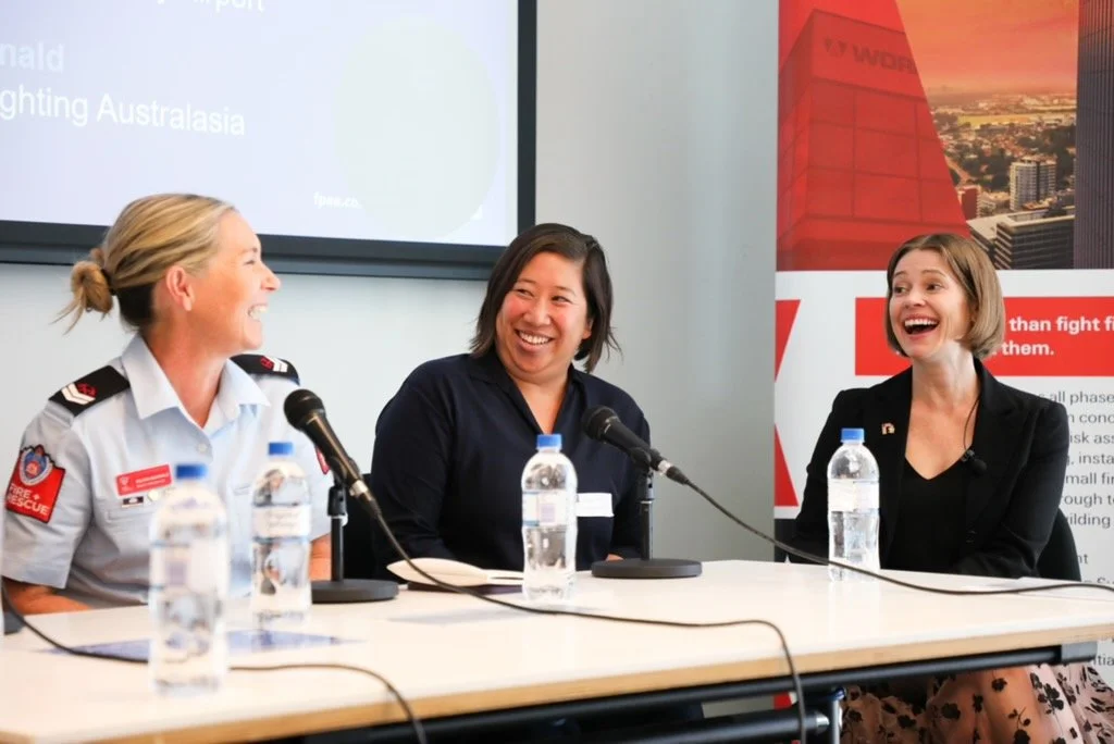 Three women sitting at a table, laughing and speaking into microphones, with water bottles in front of them, during a panel discussion or press conference.