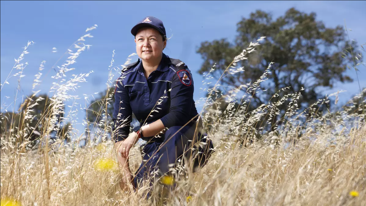 A woman dressed in a navy paramedic uniform crouching in a dry grassy field, with blue sky and trees in the background.