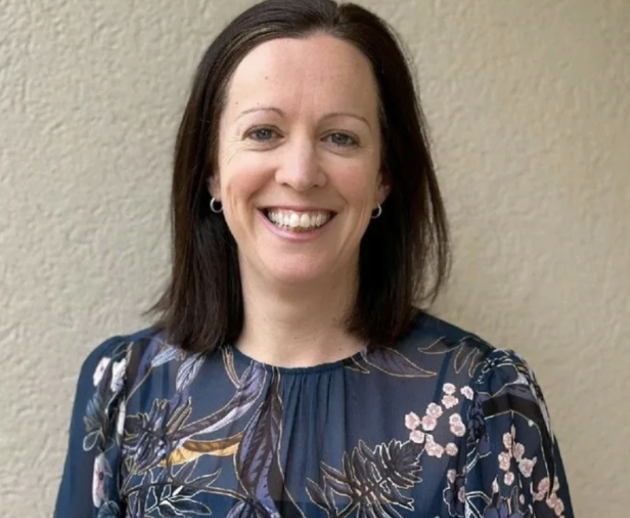 A woman with dark brown hair, smiling, wearing a blue patterned blouse, standing against a beige wall.