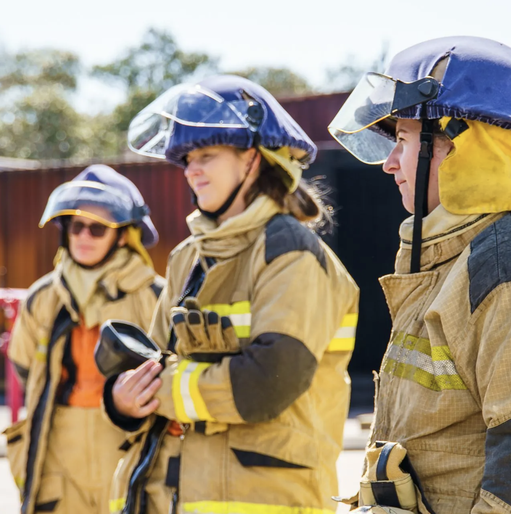Firefighters in yellow protective gear and helmets standing outdoors, with trees and a building in the background.