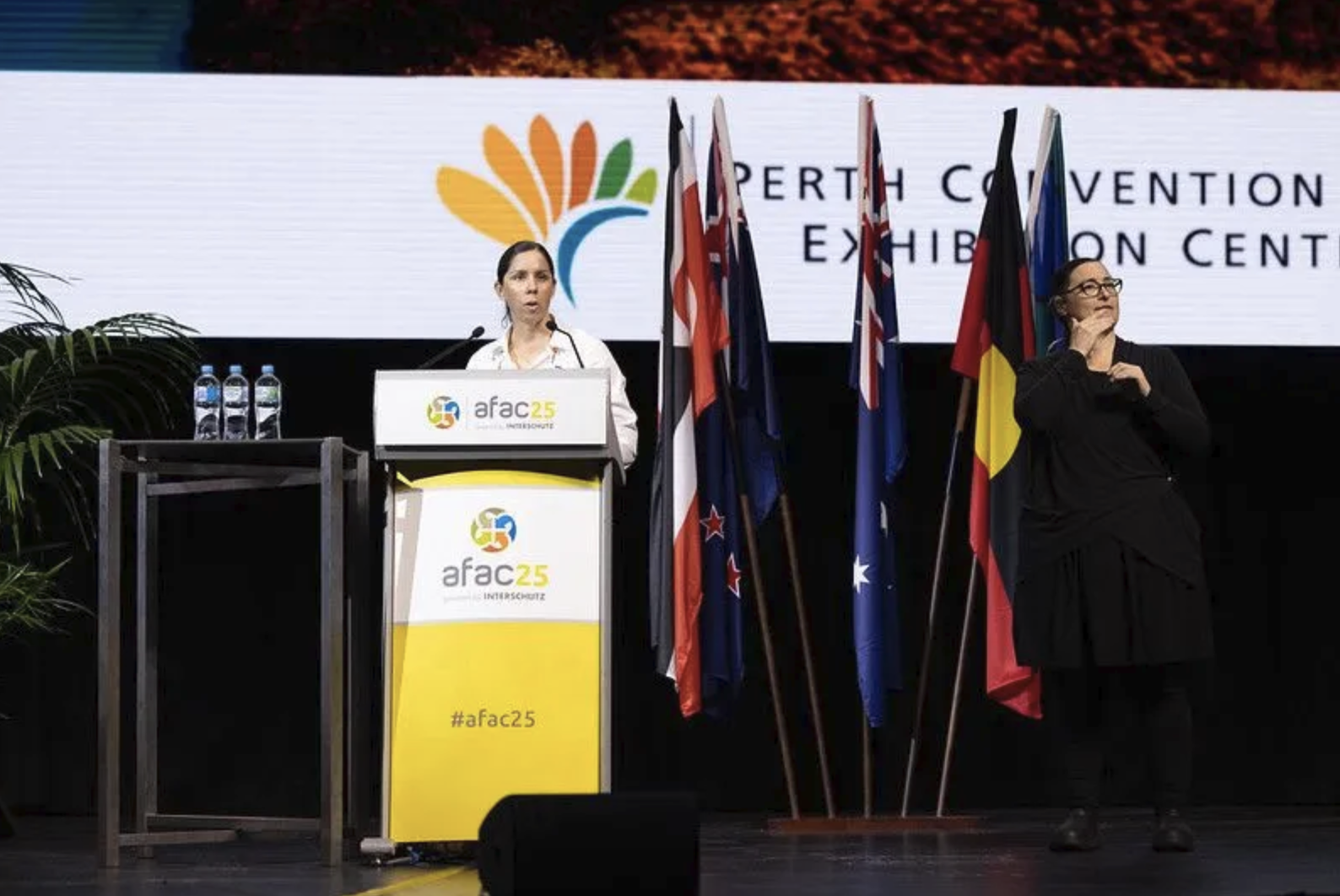 A woman speaking at a podium with bottles of water, with flags of different countries and a large screen behind her displaying the 'afac 25' logo and text about the Perth Convention Exhibition Centre.