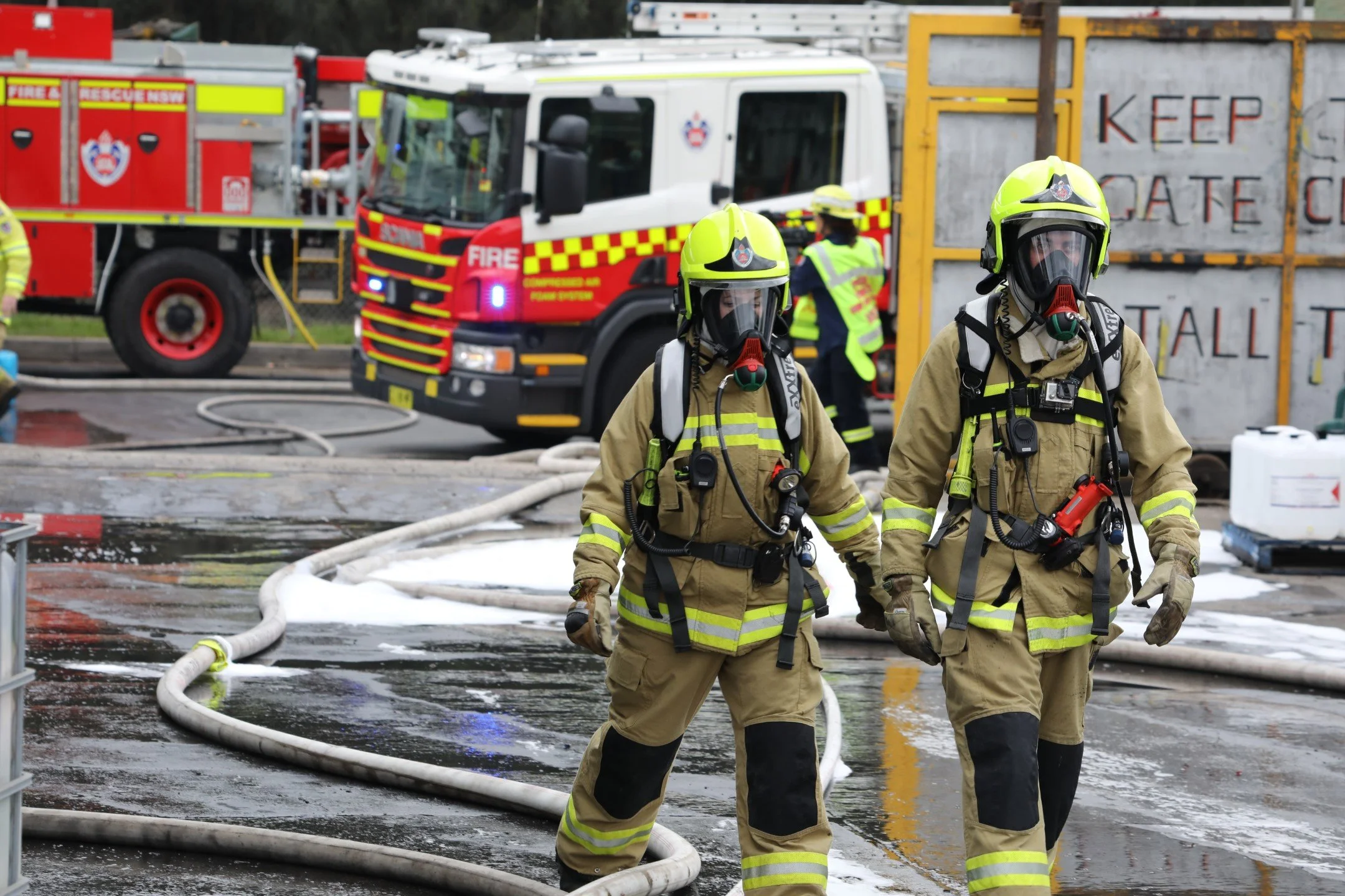 Firefighters in protective gear and helmets walking through a fire scene with fire trucks and hoses. A sign behind them reads 'Keep Gate Closed'.