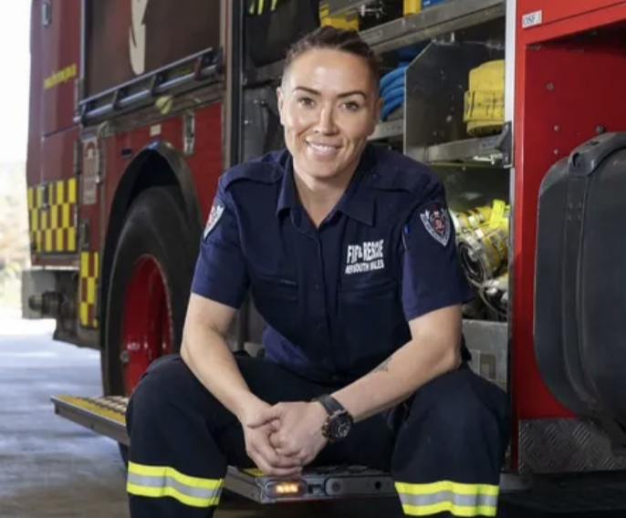 Female firefighter sitting in front of a fire truck, smiling, wearing a navy blue uniform with fire department patches, yellow reflective stripes on pants, and a wristwatch.