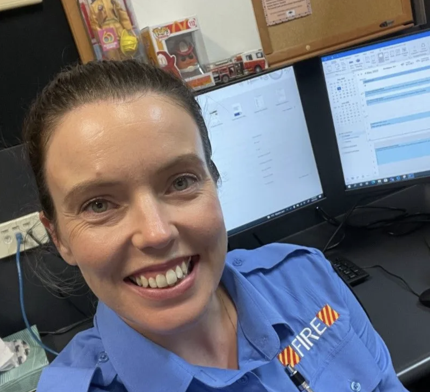 A woman in a blue firefighter uniform smiling in front of two computer monitors on a desk, with shelves of collectibles and a fire truck model in the background.