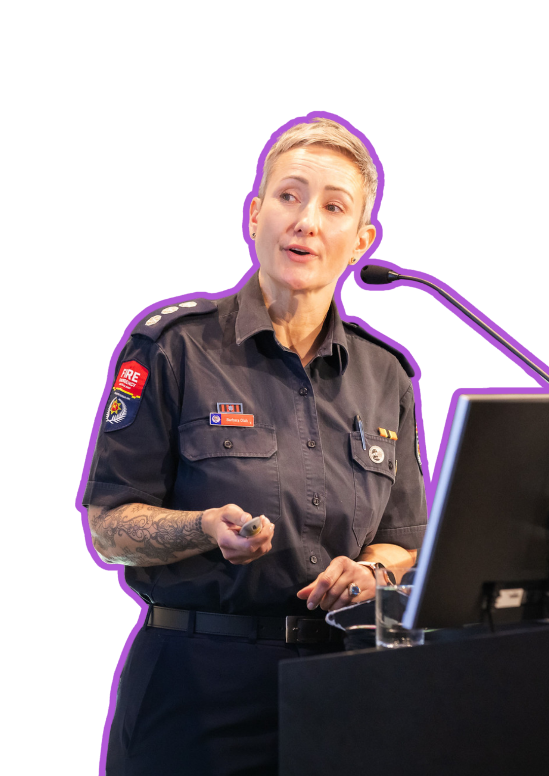 A woman with short blonde hair, wearing a black uniform with patches and badges, stands at a podium giving a presentation. She is holding a remote control in her right hand and is speaking into a microphone. The background is purple with a black table in front of her.