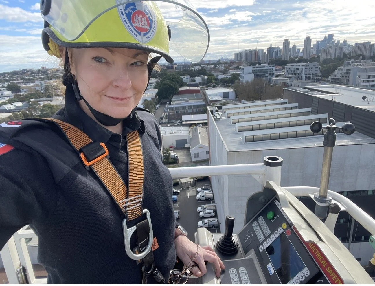 A woman in firefighter gear, wearing a helmet and harness, standing on a ladder platform high above a cityscape with buildings and a partly cloudy sky.