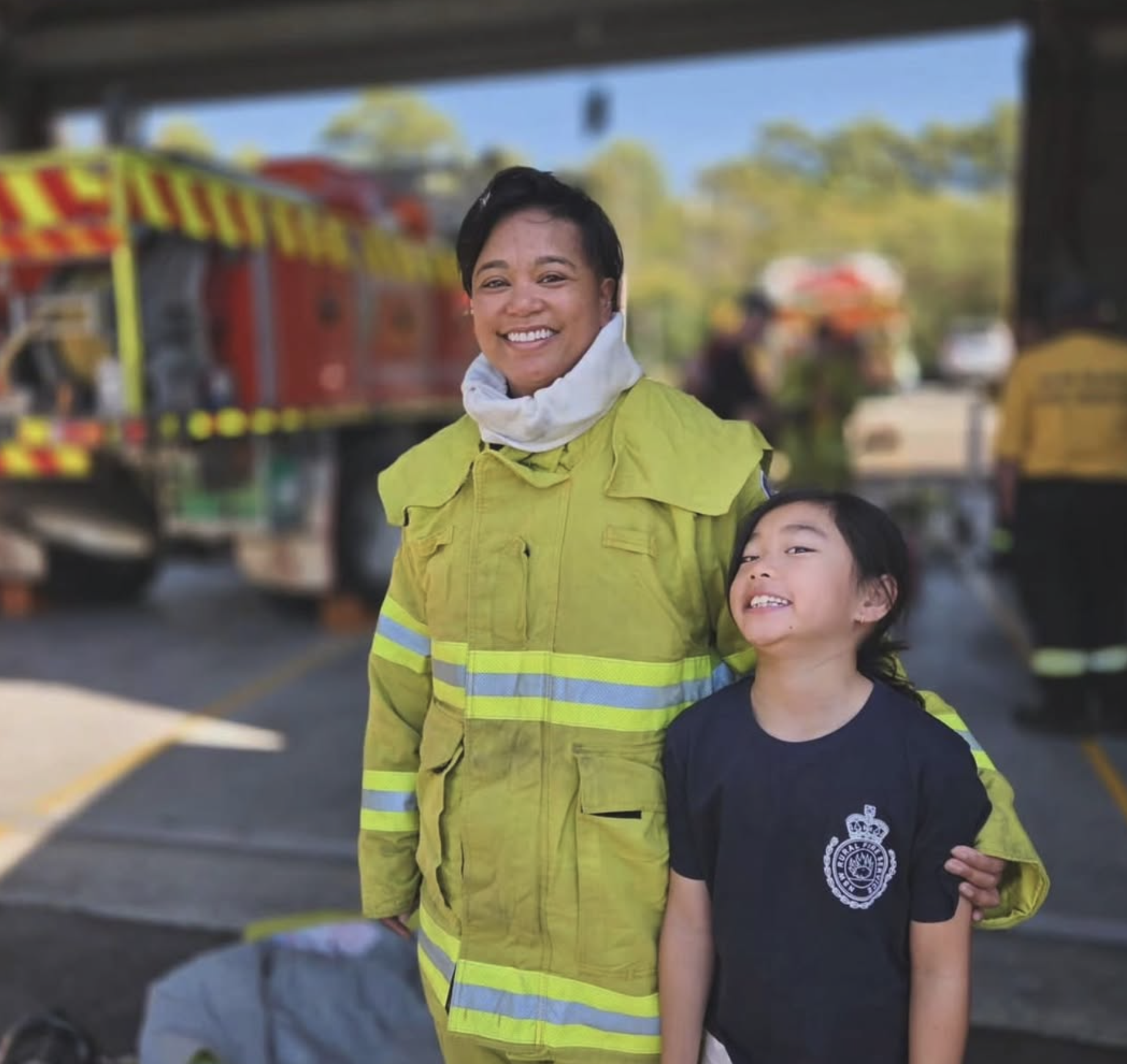 A woman with short dark hair wearing a high-visibility yellow safety jacket and a white face mask around her neck, smiling for the camera, standing next to a young girl with long dark hair wearing a dark T-shirt with a crown emblem. They are outdoors with trucks and trees in the background.