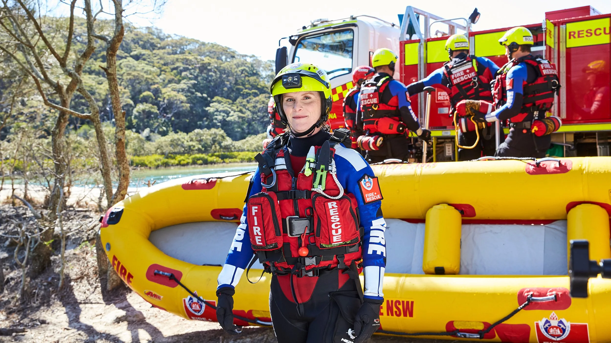 A female emergency rescue worker standing in front of a yellow rescue boat, wearing a yellow helmet, red rescue vest, and blue and white uniform, with other rescue team members and a rescue vehicle in the background.