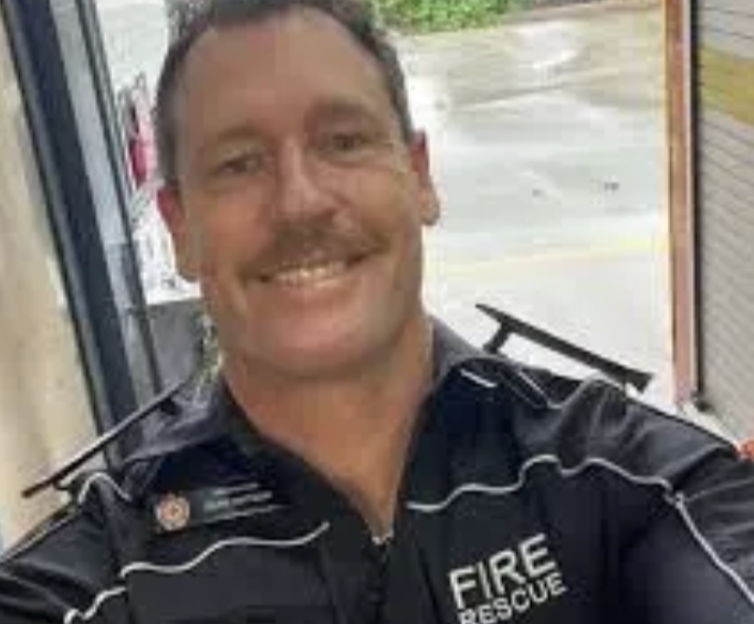 A smiling man in a fire rescue uniform taking a selfie indoors with a glass door and outdoor parking lot in background.