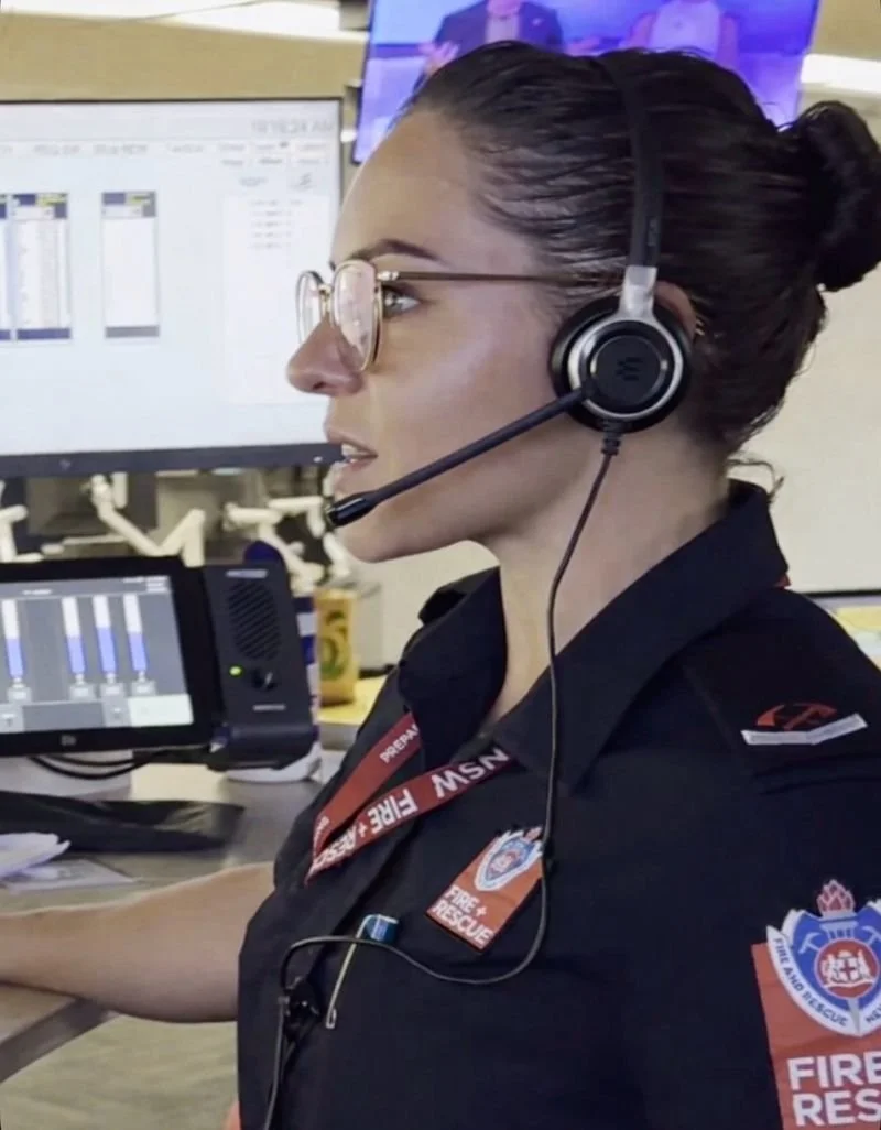 A female firefighter at a desk wearing a headset, glasses, and a uniform with fire department insignia, working with multiple computer monitors.