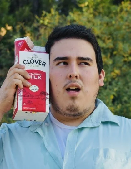 A man holding a carton of Clover Sonoma Vitamin D Milk to his ear, making a silly face outdoors with trees in the background.