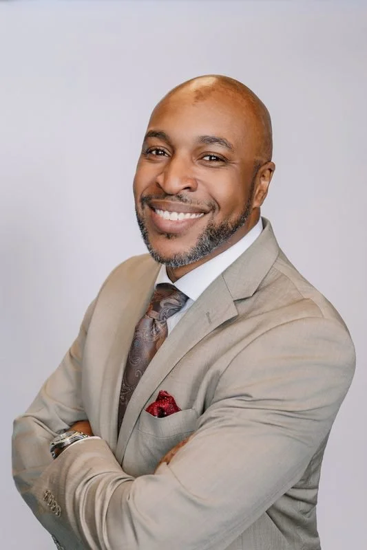 A smiling African American man in a beige suit, white shirt, and patterned tie, with a red pocket square, standing with crossed arms against a light gray background.