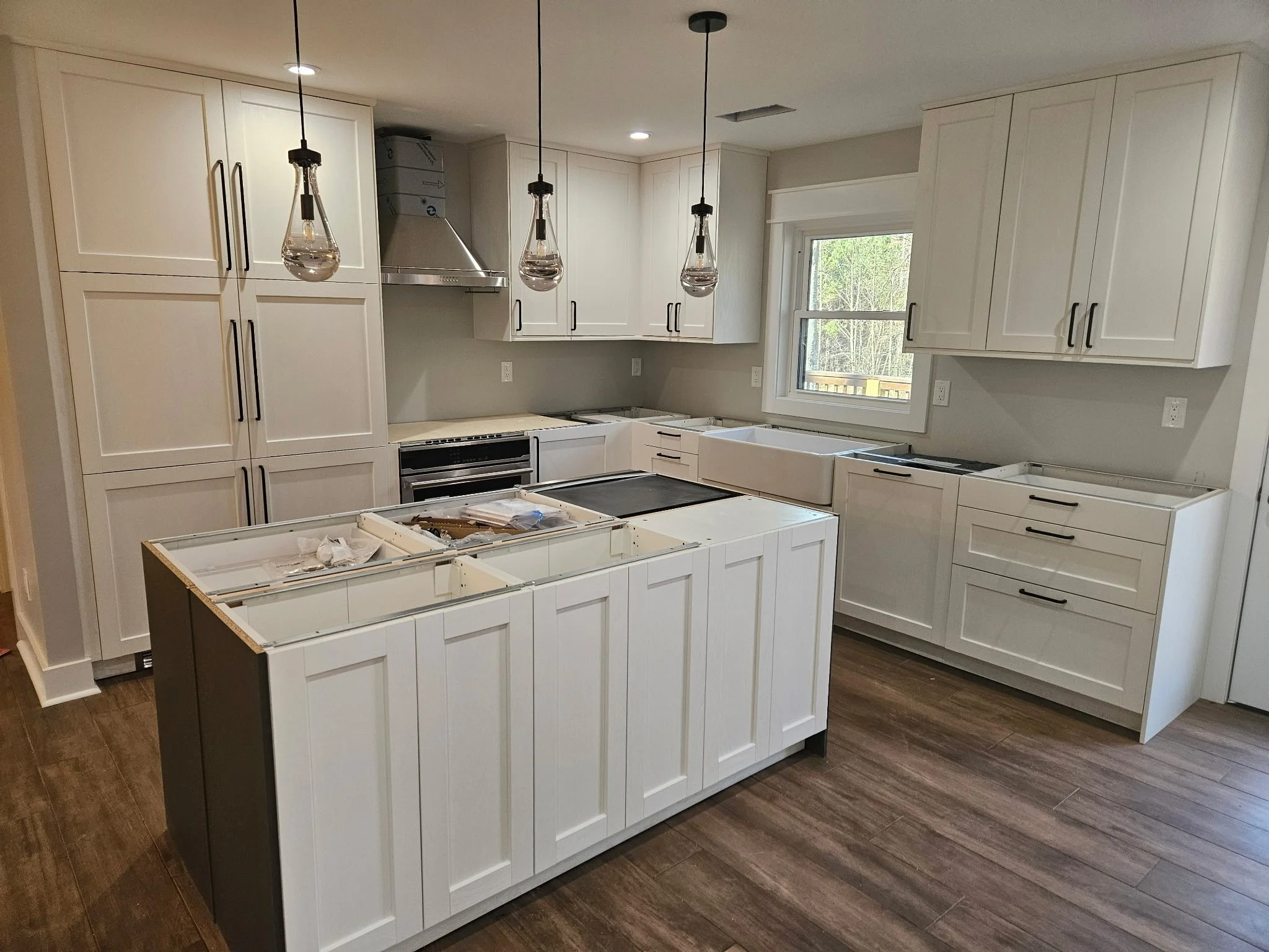 An unfinished kitchen with white cabinetry, a dark wood floor, and three pendant lights hanging above a kitchen island. The island has no countertop yet, and there are boxes and packaging on top.