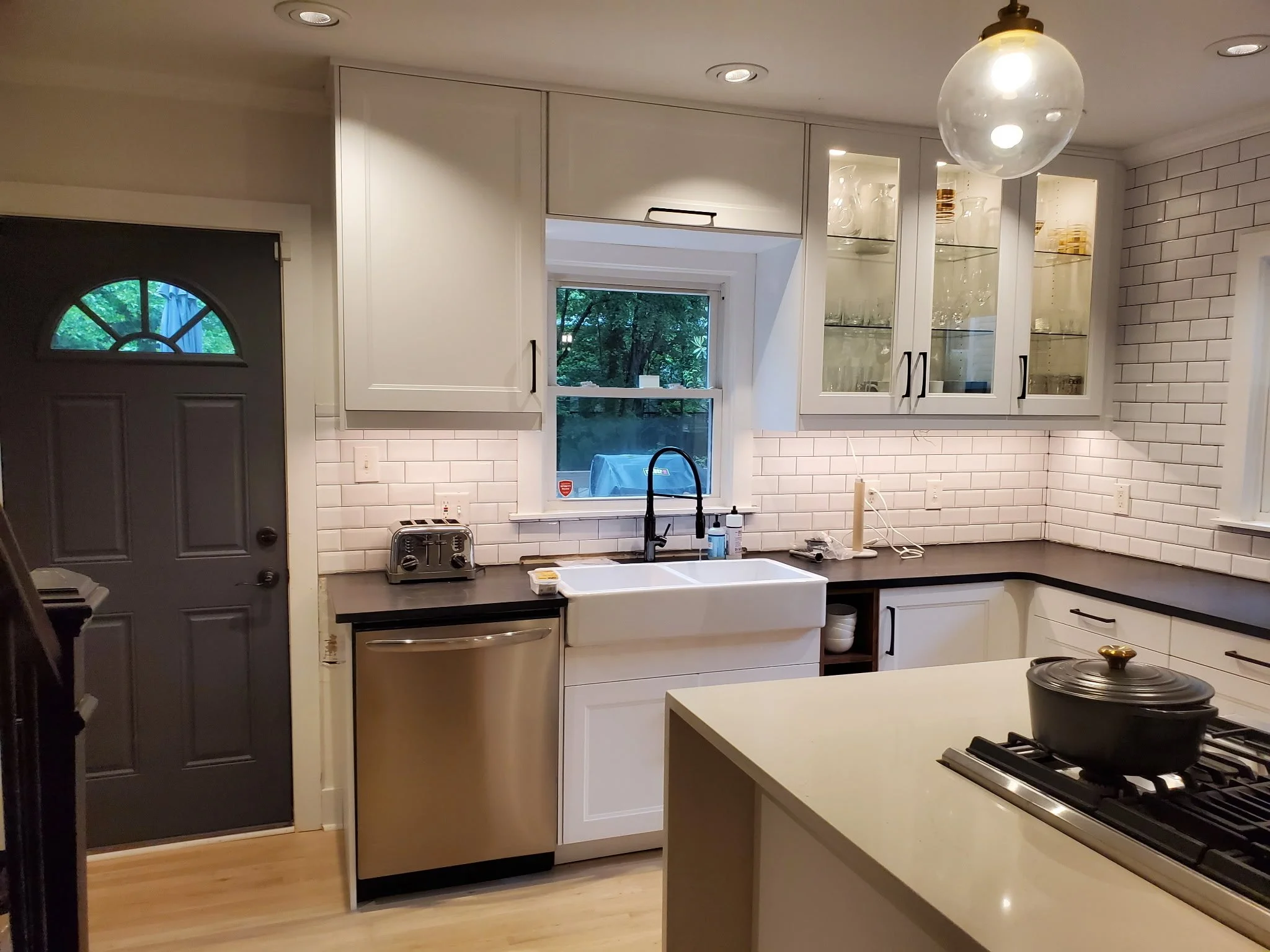 Kitchen with white cabinets, a black countertop, and a white subway tile backsplash. There's a window above the sink showing trees outside, a black faucet, and a stove with a black pot. A hanging light fixture is above, and to the left is a gray door with a half-moon window.