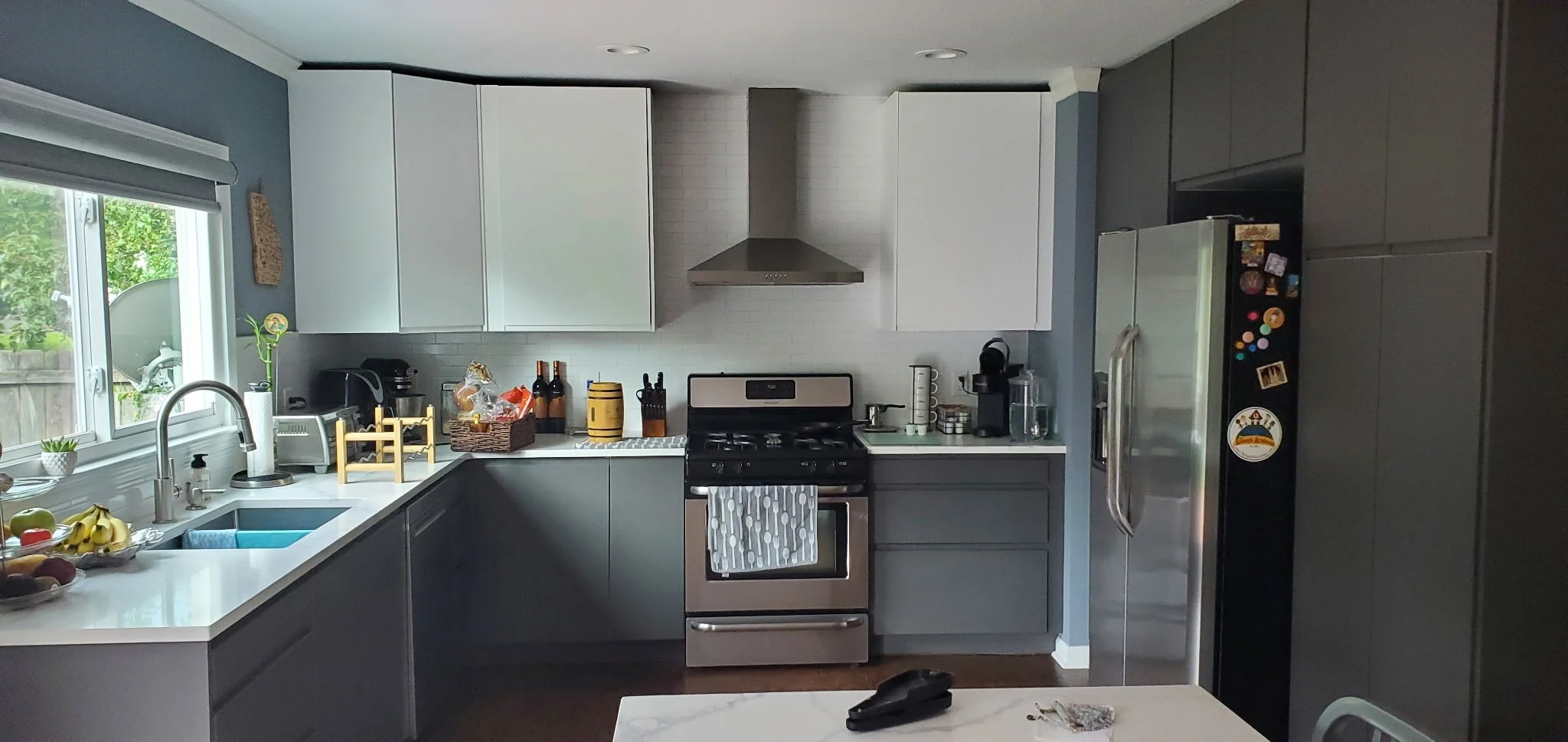 A modern kitchen with a white countertop, gray cabinets, and a window with a view of greenery. The kitchen includes a stainless steel stove, black refrigerator with magnets, and various kitchen appliances and utensils on the counter.