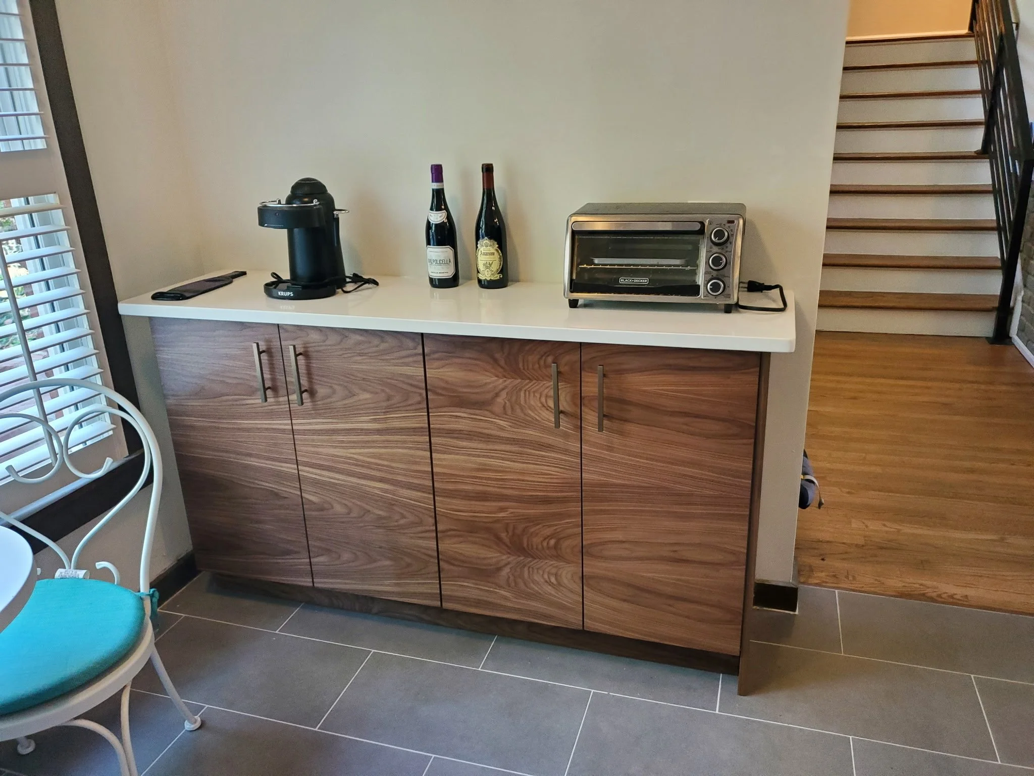 A kitchen or dining area showing a white countertop with a coffee machine, two bottles of wine, and a toaster oven on it, with a wood cabinet below, next to a staircase with wooden steps and a black railing.