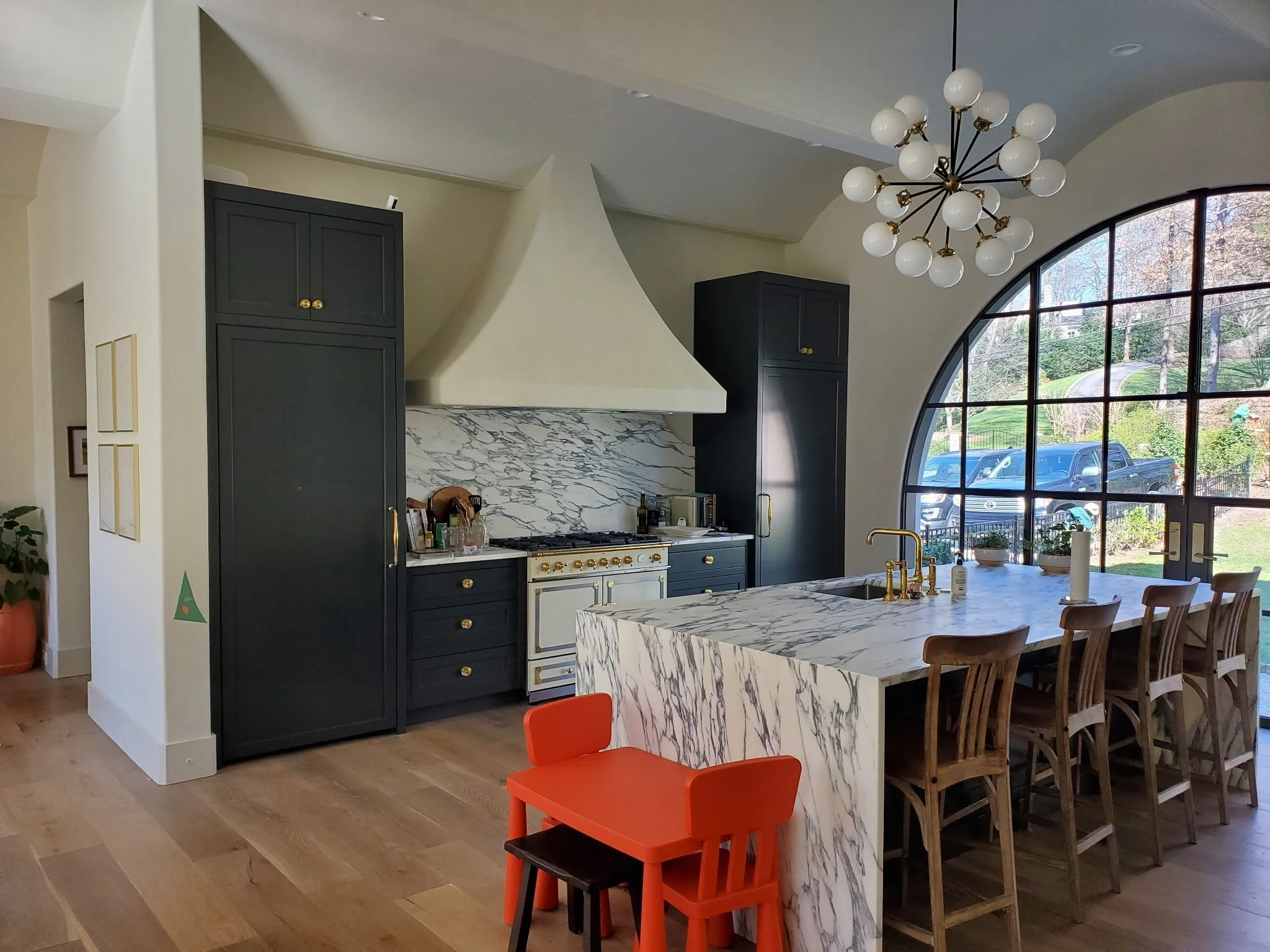 Interior view of a modern kitchen and dining area featuring dark cabinets, a marble kitchen island, a large arched window, and a chandelier with multiple white globes.