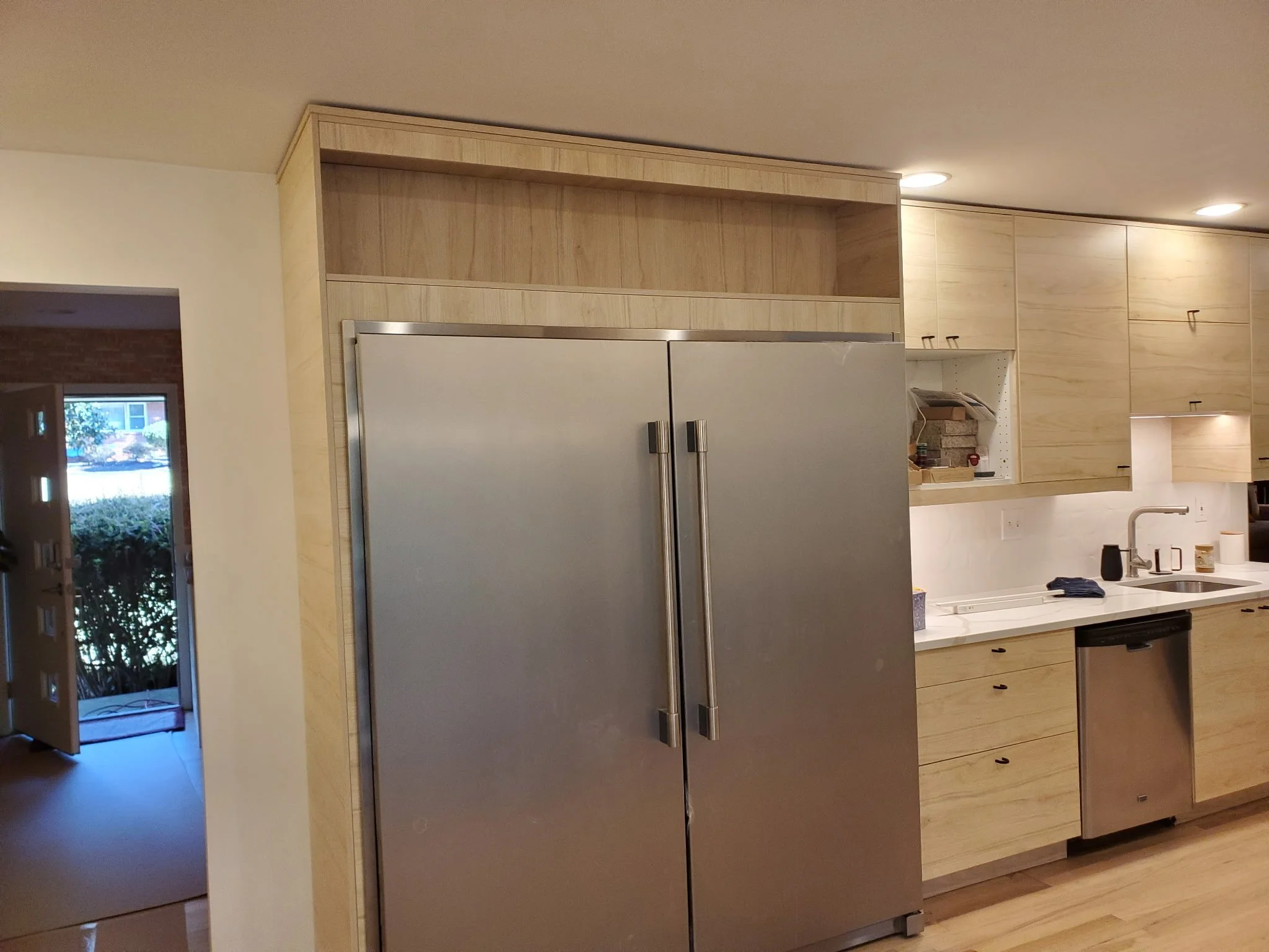 A modern kitchen with wooden cabinets, a stainless steel refrigerator, and a white countertop. There is an opening to the left showing a door to the outside with trees and a building visible through the glass.