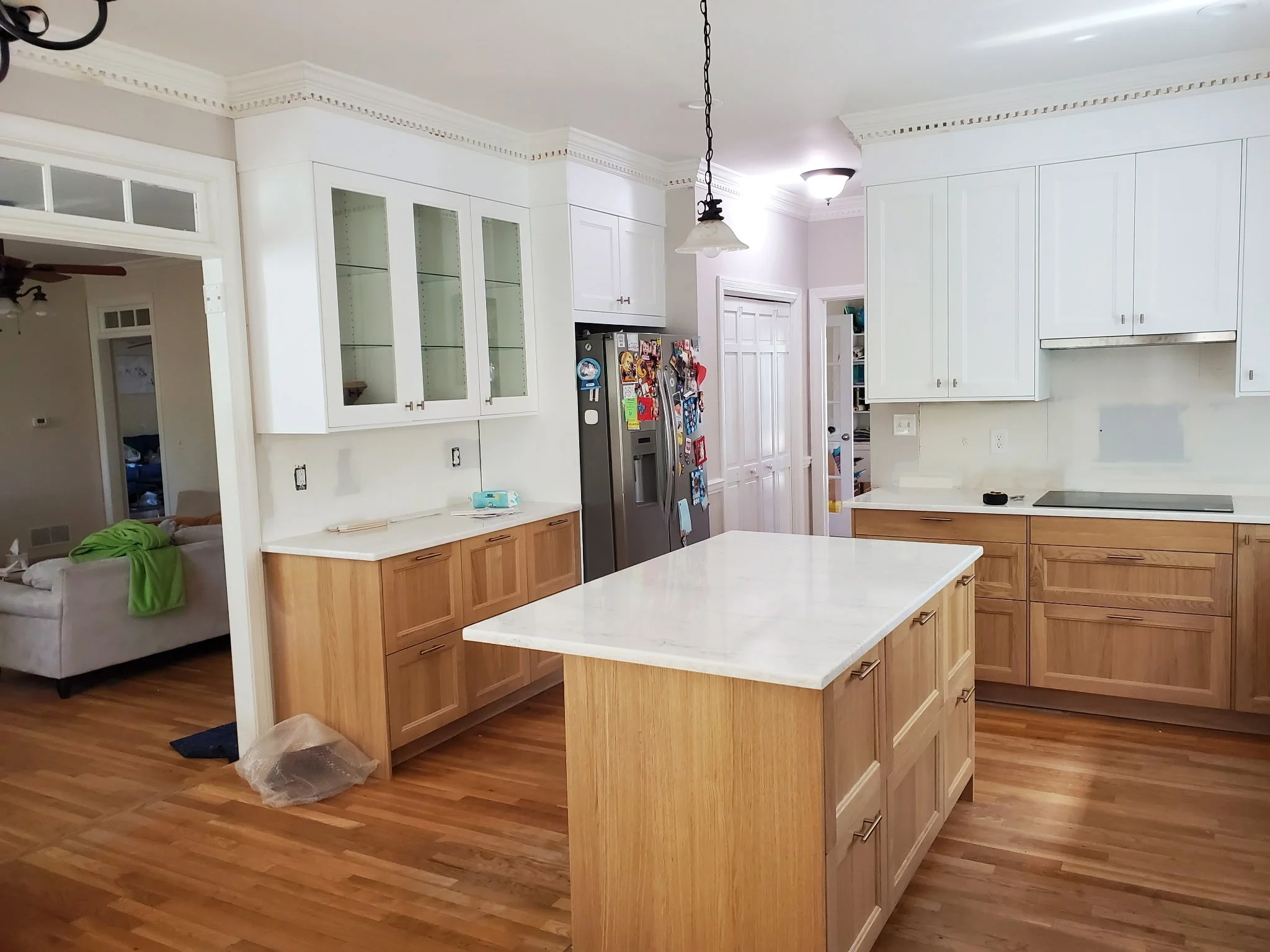 A kitchen with white upper cabinets and wooden lower cabinets, a white countertop, a stainless steel refrigerator with magnets, and a kitchen island with a white countertop and wooden drawers. The floor is wood, and there is a doorway leading to another room.