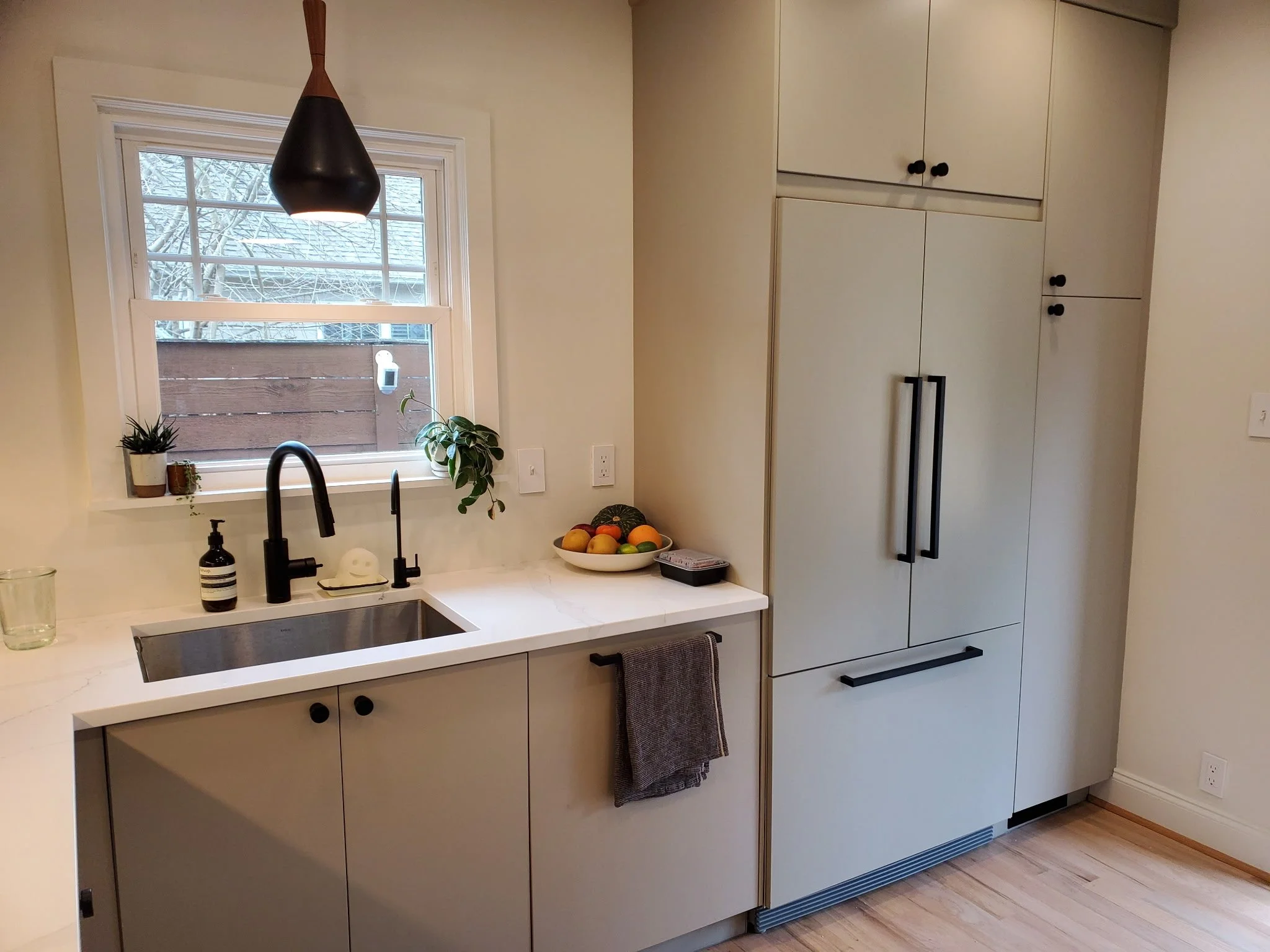 A modern kitchen with white cabinets, a white countertop, a black faucet, a hanging black pendant light, a window with a snow-covered tree outside, and a bowl of mixed fruits on the counter.