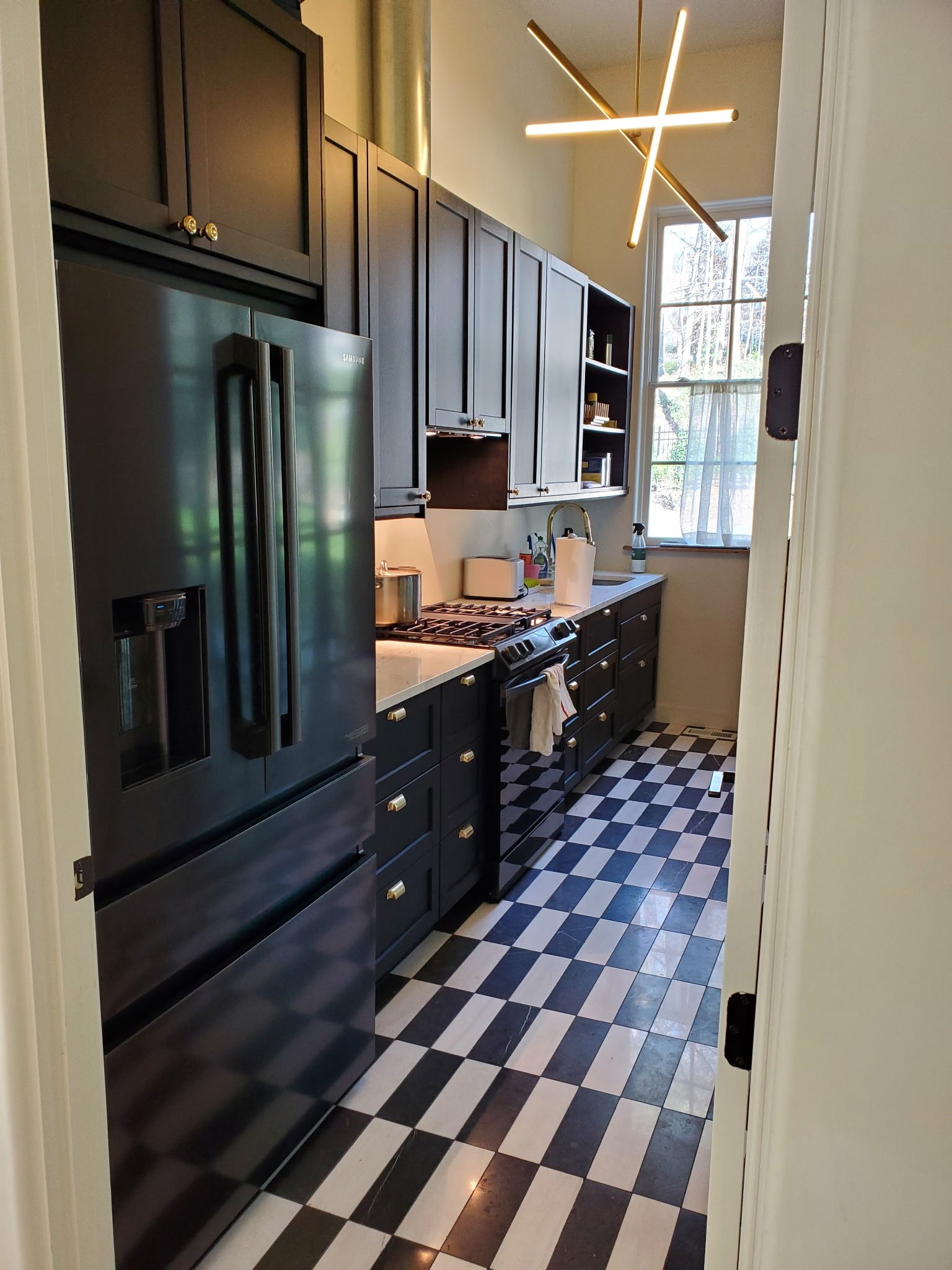 A narrow kitchen with black and white checkered floor tiles, dark cabinetry, a stainless steel refrigerator, and a large window with curtains letting in natural light, along with a modern ceiling light fixture.