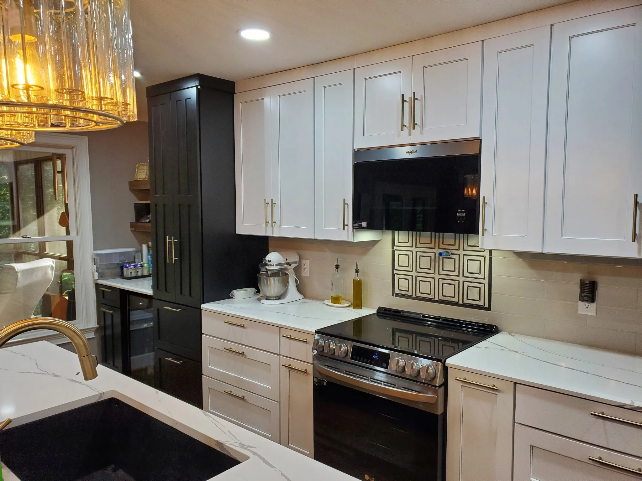A modern kitchen with white cabinets, a black oven, a white countertop, a gold faucet, and a black and gold backsplash. There is a round, gold chandelier above the island and a sliding glass door on the left side.